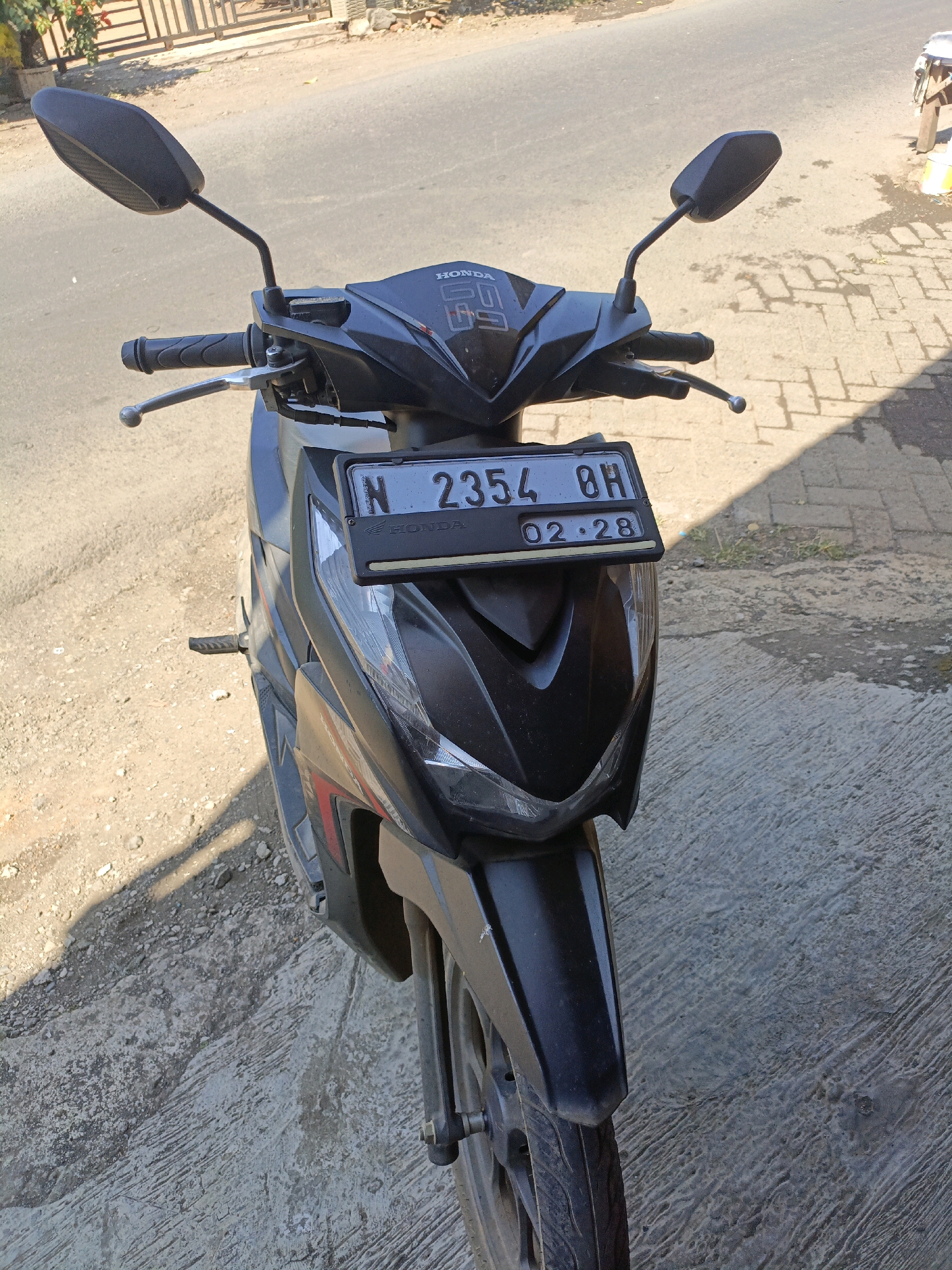 A black Honda scooter is parked on a paved surface, likely a sidewalk or street, with a dirt and gravel area to its left. The scooter's license plate reads "N 2354 BH" and below that "02-28". The scooter is facing forward, with its handlebars, mirrors, and front fender clearly visible. The background shows a road and a fence with some foliage behind it. The scene appears to be outdoors in a sunny environment, with distinct shadows cast on the ground. The location is identified as Betek, Indonesia. There are no people or animals visible in the image.