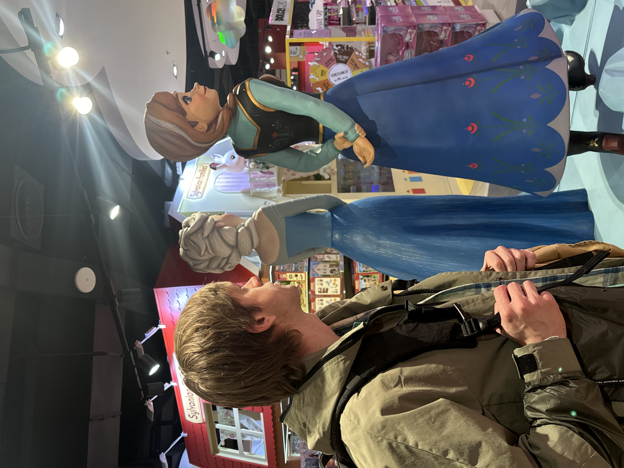 The image shows a man looking at statues of the characters Anna and Elsa from the Disney film "Frozen." The location is the Central Children's Store in Moscow, Russia, as indicated by visible signage and the store's layout. The man is wearing a green jacket and backpack and is facing upwards towards the statues. The statues are large and detailed, depicting Anna in her green and black outfit and Elsa in her blue dress. Background details include store shelves filled with toys and other merchandise. The lighting is bright, with visible spotlights.