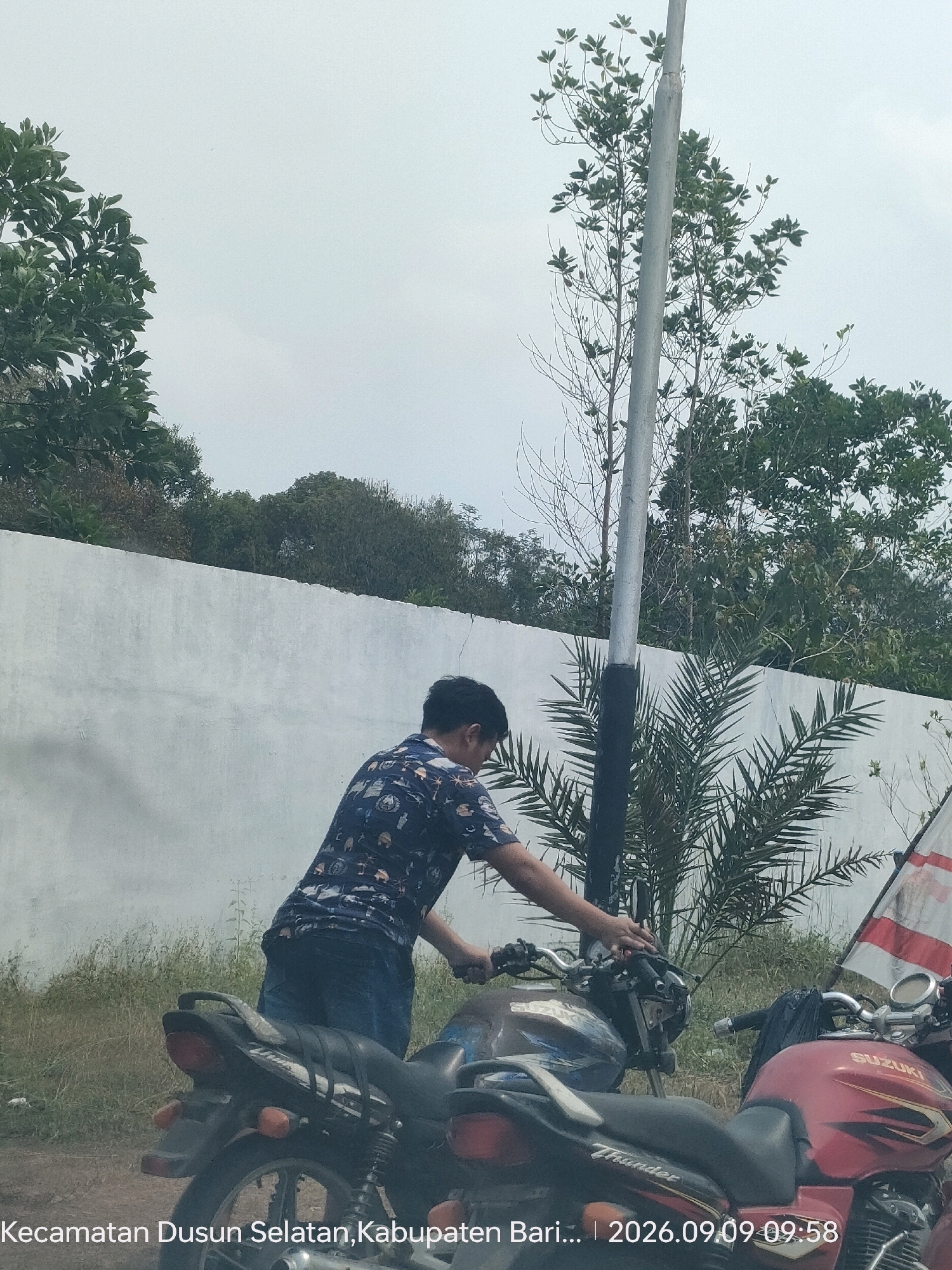 In Sababilah, Indonesia, a young man wearing a dark patterned shirt and blue jeans is seen leaning over a motorcycle. The motorcycle is black with some blue accents and has the word "Thunder" written on its side. Another motorcycle, red with "Suzuki" branding, is parked next to it. Behind the man and motorcycles is a white wall topped with lush green trees. A tall, slender pole, possibly a lamppost, stands prominently in the foreground, partially obscured by the man and the trees. The sky is overcast, and the overall setting appears to be outdoors with some vegetation and a dirt or gravel path. The bottom of the image displays text that reads "Kecamatan Dusun Selatan, Kabupaten Bari... | 2026.09.09 09:58".