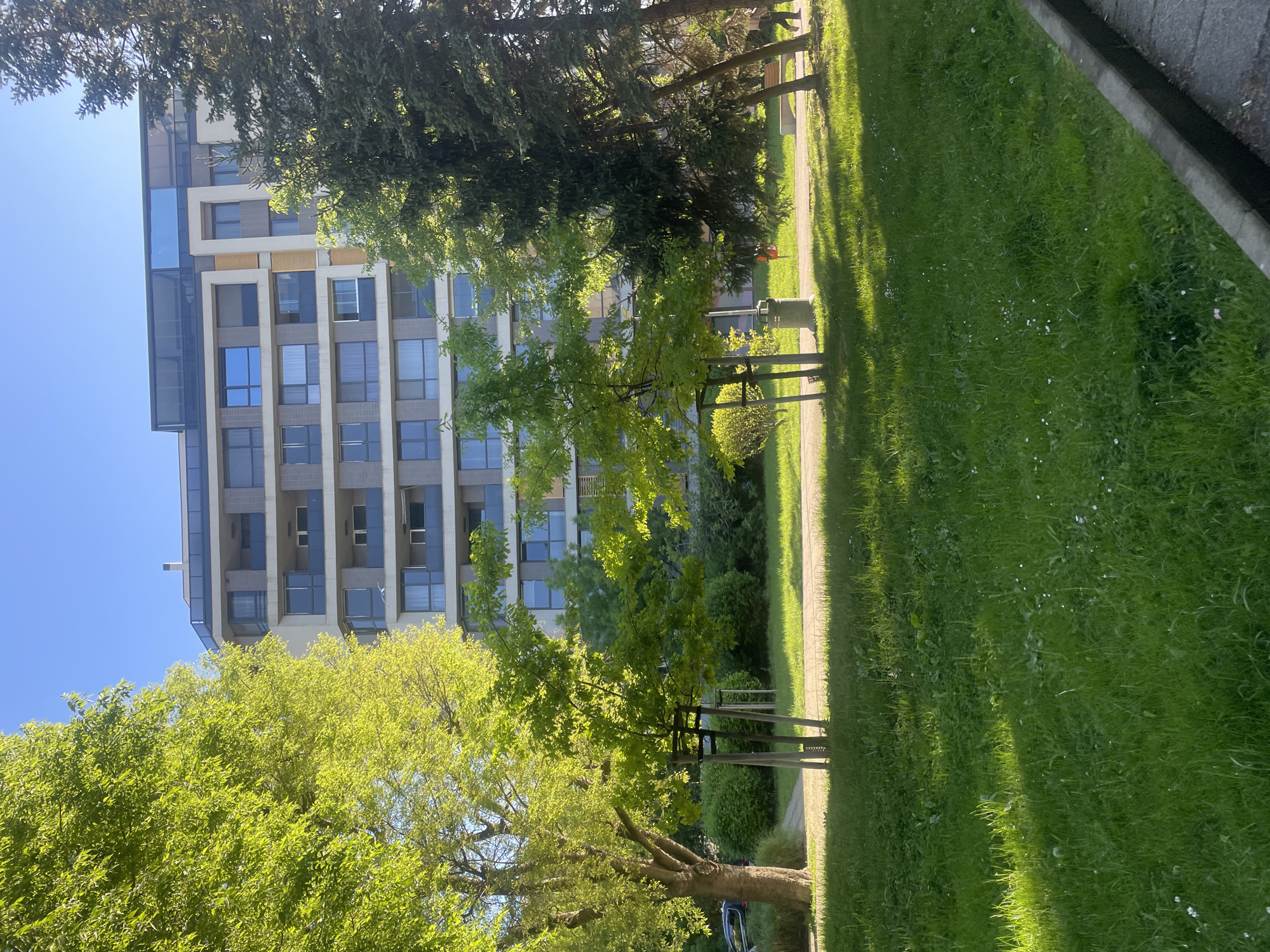The image shows a multi-story building with a modern facade, likely residential apartments, in Burgas, Bulgaria. The building has numerous windows and some balconies. In the foreground, there is green grass and several trees. Some of the trees are young and have support structures around them. There is a paved pathway between the grass and the trees. The sky is blue and clear, indicating a sunny day. A small glimpse of a vehicle is visible.