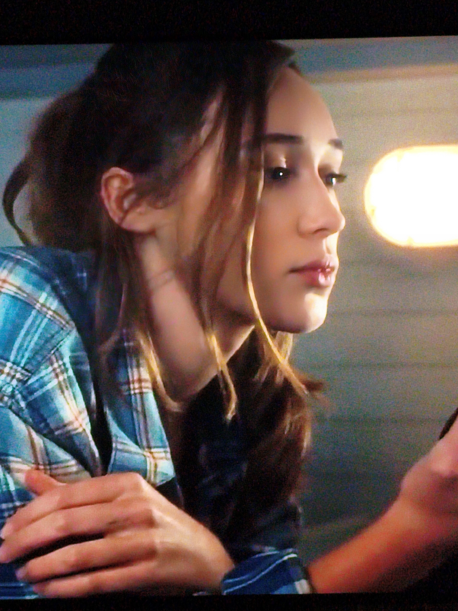 The image is a close-up shot of a young woman with brown hair, wearing a blue and white plaid shirt. She is looking to her right, with her head tilted slightly. Her right hand is resting on her chest, with her fingers interlaced. A blurred background suggests an indoor setting, possibly a room with a warm, glowing light source on the right side of the frame. The woman's facial features, including her eyes, nose, and lips, are visible. The overall lighting is dim, with the main light source illuminating the woman's profile. There is no indication of the location being in Vladivostok, Russia.