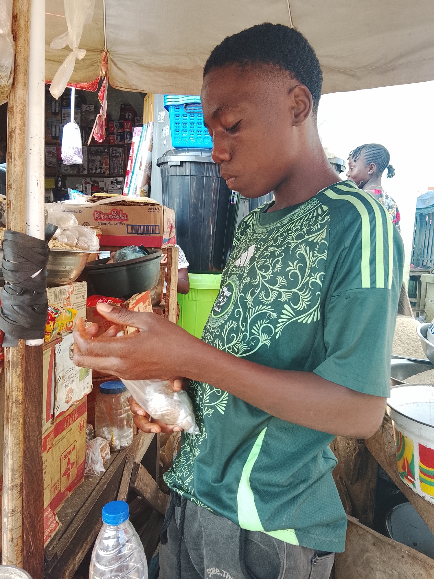 A young man, appearing to be in his late teens or early twenties, is seen standing at a market stall in Akwanga, Nigeria. He is wearing a dark green t-shirt with a light green patterned design and light green stripes on the sleeves. His hair is short and dark. He is holding a small, clear plastic bag containing what appears to be food items, and is looking down at it, seemingly in the process of examining or preparing its contents. His hands are open, and he is carefully manipulating the bag.
The market stall is constructed from wood and appears to be set up outdoors, with a light-colored tarp providing shade overhead. Various items are visible on the shelves of the stall, including boxes of "Kremela" and "Maggi" products, plastic containers, bowls, and bags of what might be grains or other dry goods. A blue plastic basket is stacked on top of a large dark container. A bottle of water with a blue cap is also visible in the foreground. In the background, another person, a woman with braided hair, is partially visible, looking in the same general direction as the young man. The overall impression is of a busy, informal marketplace setting.