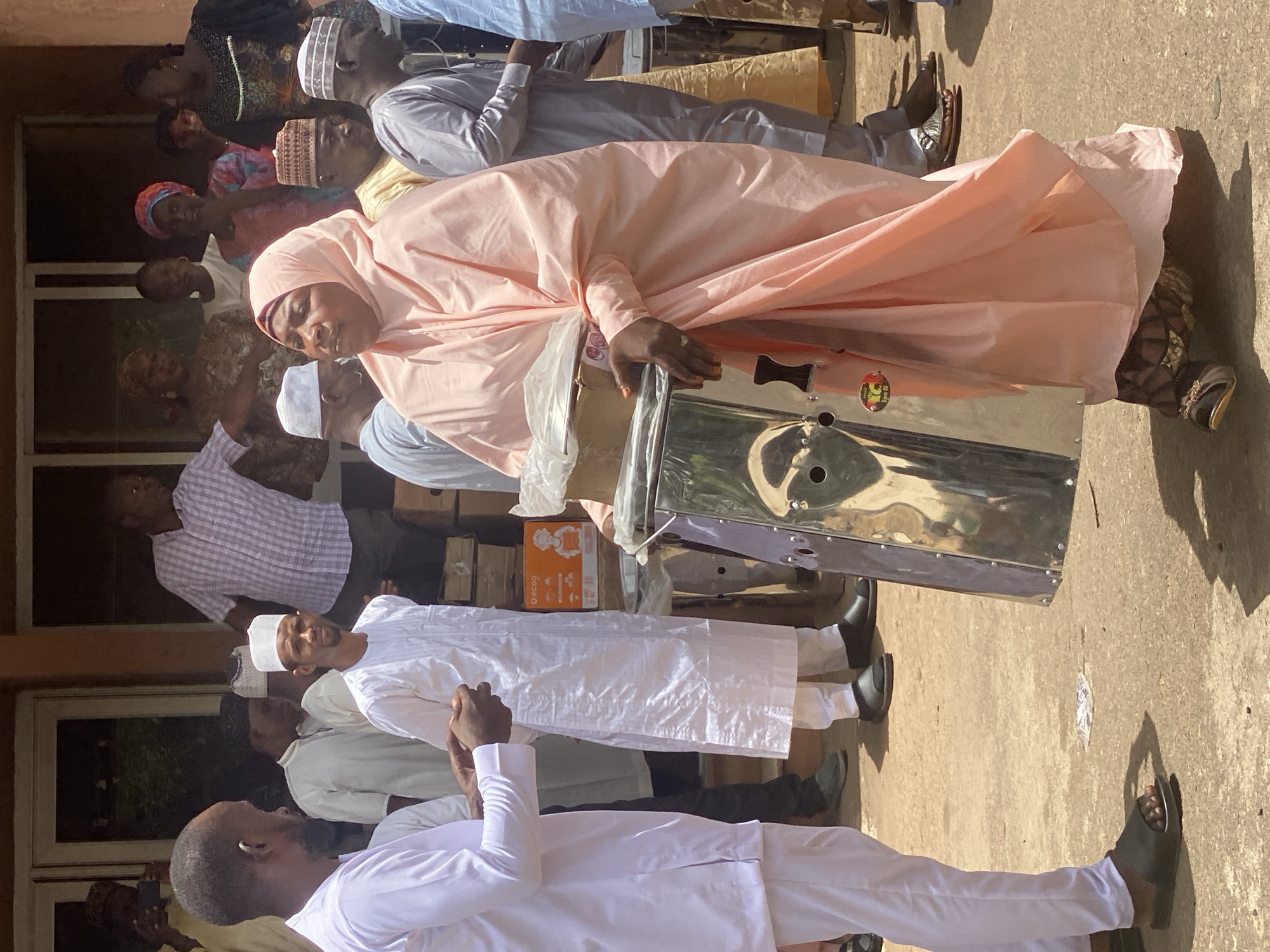 In Unguwar Rimi, Nigeria, a group of people is gathered outdoors. A woman in a light orange hijab is carrying a metallic object, possibly a machine or appliance. Several men are dressed in white traditional garments and caps. Others are wearing casual attire. The environment appears to be a paved area in front of a building, with additional people visible in the background. There's evidence of boxes or packaging near the metallic object. The scene is brightly lit with sunlight, casting shadows.