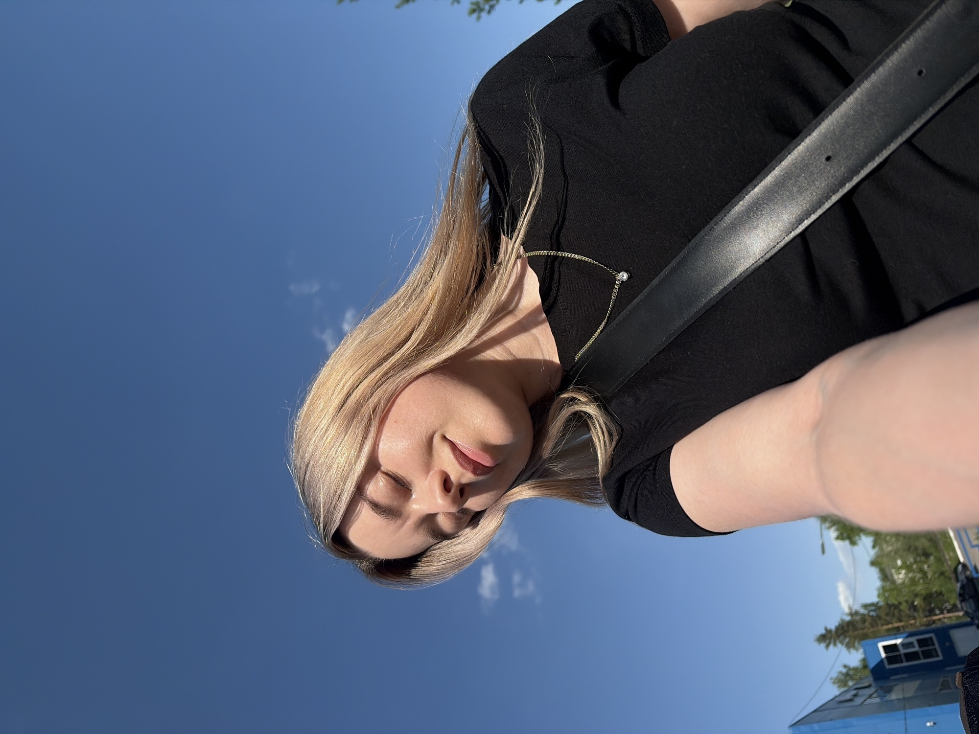 A woman with blonde hair, wearing a black top and a black belt across her chest, is taking a selfie against a clear blue sky. Her eyes are closed, and she appears to be tilting her head. The background shows parts of a city landscape. A blue trailer with windows is visible, along with trees and a street lamp. Based on its location in Chaikovsky, Russia, the image depicts a person taking a selfie outdoors, possibly in a park or urban area.