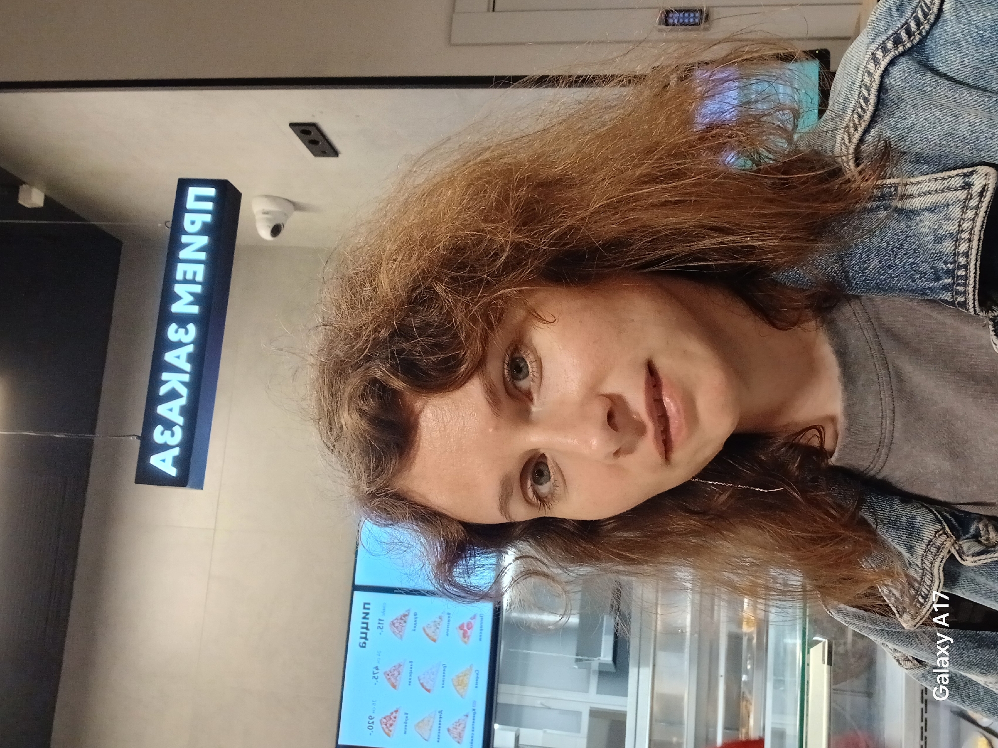 The image shows a woman with light brown, curly hair, wearing a gray shirt and a denim jacket. She is standing indoors, possibly in a pizzeria in Yekaterinburg, Russia. In the background, there is a blue sign with white letters that says "АЗАКАЯ МЗNЭП," as well as a menu screen displaying images and prices of pizzas. A security camera is visible near the sign. A Galaxy A17 phone is visible in the bottom right corner of the frame.