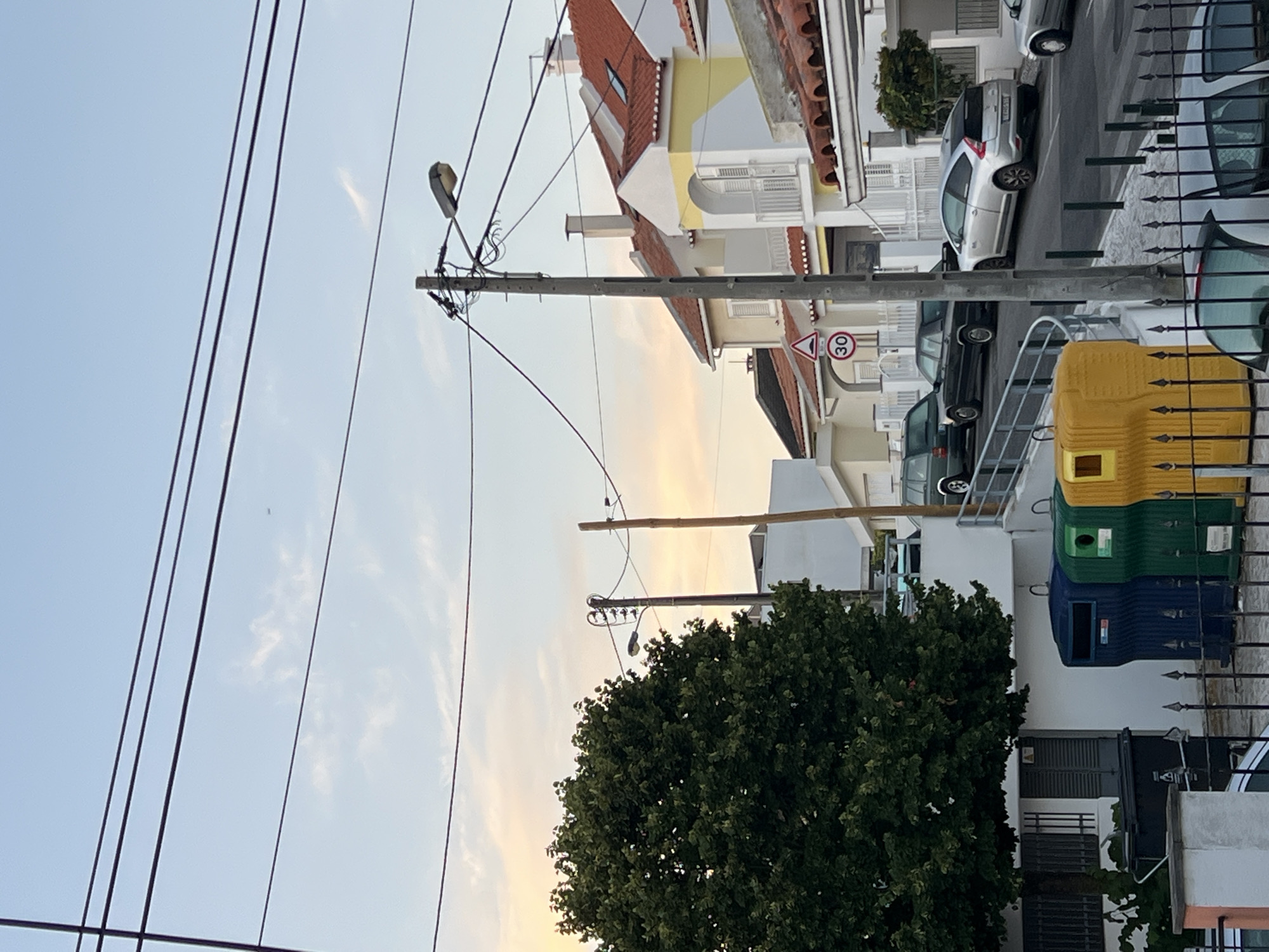 The image depicts a street scene in Cascais, Portugal, likely during the late afternoon or early evening, as indicated by the warm, soft light illuminating the sky. The foreground is dominated by electrical poles and wires, a large green tree on the left, and three colorful recycling bins (blue, green, and yellow) positioned next to a dark-colored trash bin. A black metal fence runs along the bottom of the frame, with parts of vehicles visible behind it.
The mid-ground reveals a street with parked cars. A silver hatchback is prominently featured on the right side of the street, alongside other vehicles, including a dark-colored sedan and a dark green SUV. The street is lined with residential buildings characterized by their red-tiled roofs and white or pale yellow facades, some featuring balconies and arched windows. A speed limit sign indicating 30 km/h is visible on the left side of the street.
The background consists of a pale blue sky with scattered clouds tinged with orange and yellow hues, suggesting the setting sun. The overall impression is of a typical suburban or residential street in a coastal town.