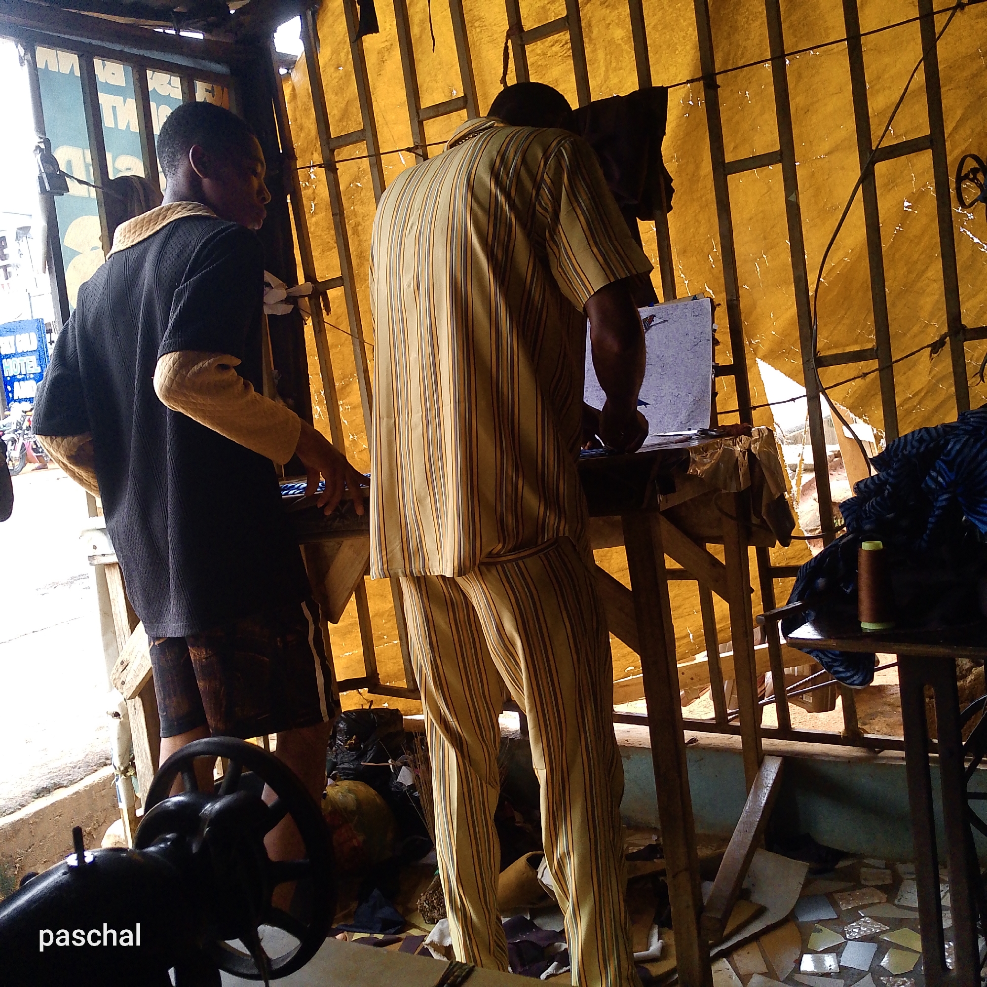 The image depicts a scene at a tailor's shop in Ukwu-Abwa, Nigeria. Two men are present. One man wears a black shirt with tan sleeves and dark shorts. He is standing at a work table. The other man is clad in a matching striped yellow and tan shirt and pants. He stands at another work table writing on a piece of paper. Both men appear to be focused on their work. The shop's interior is simple, with a makeshift wooden structure forming the walls and a yellow tarpaulin roof. A sewing machine is in the lower left corner. The floor shows discarded scraps. Outside, a street is visible.