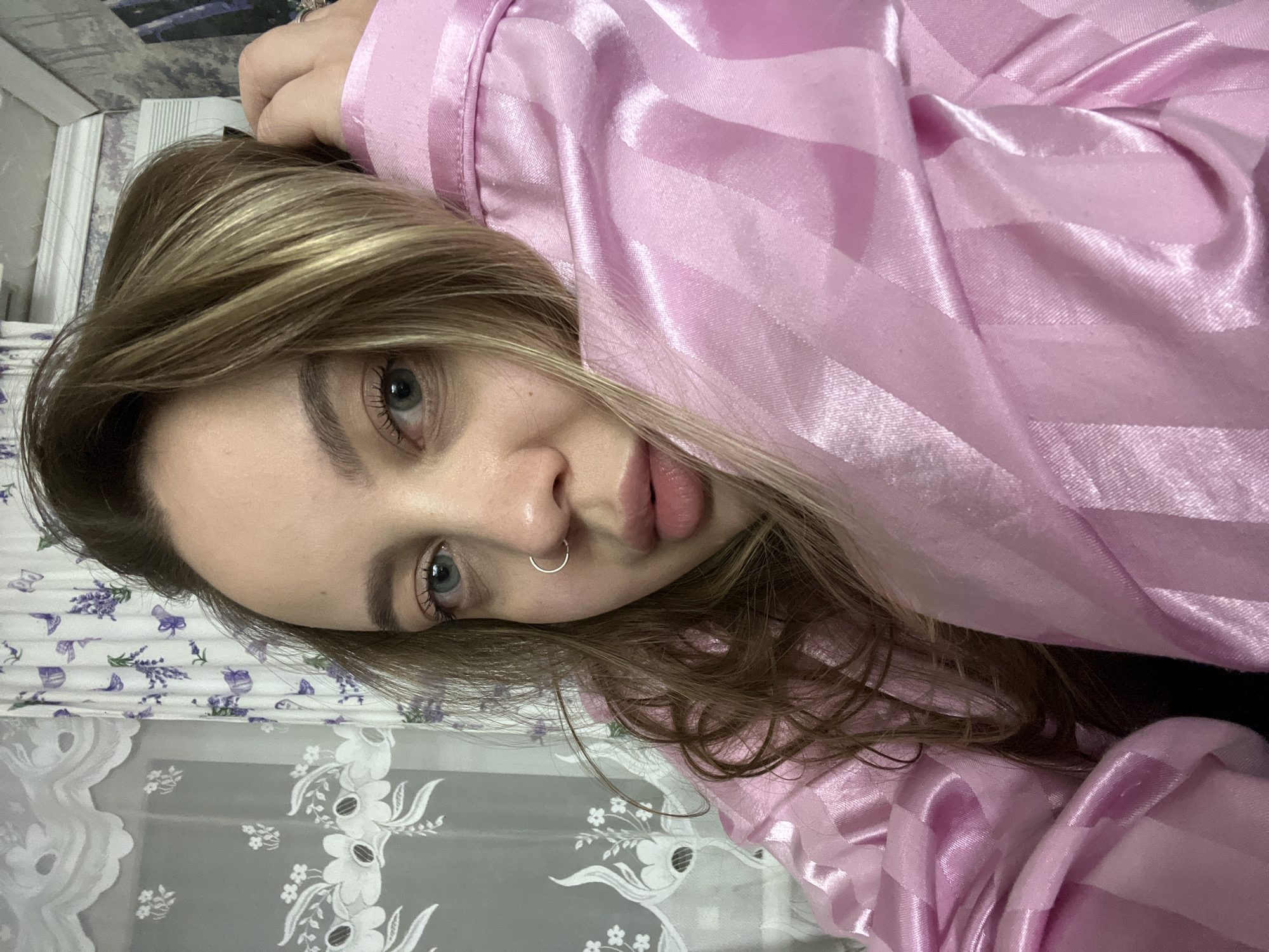 A woman with blonde hair and a nose ring is lying down, wearing a pink striped shirt. She is indoors with floral patterned bedding and curtains. The location in Novopokrovskaya, Russia is not discernible from the image.