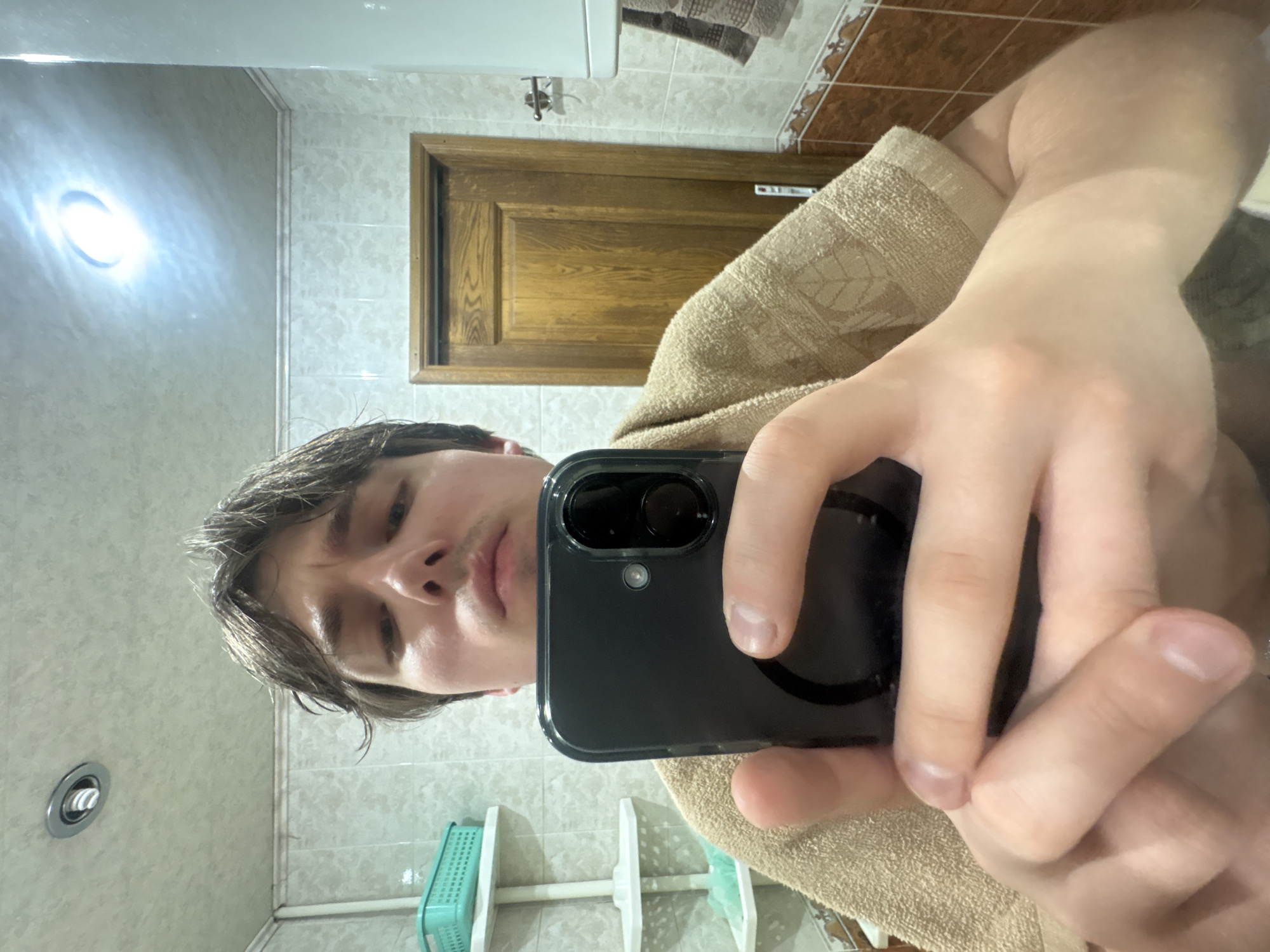 A young man with light brown hair is taking a selfie in a bathroom. He is wearing a light brown towel around his shoulders. He is holding a black smartphone with his right hand, and his left hand is visible in the foreground. The bathroom appears to have tiled walls and a wooden door in the background. There are shelves with a green basket on the left side of the image. A light fixture is visible on the ceiling. The scene is likely indoors, possibly in a residential building in Dnipro, Ukraine, given the context provided.