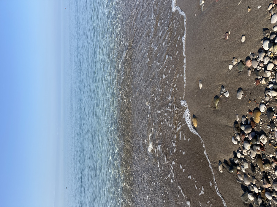 The image depicts a tranquil coastal scene at Šušanj, Montenegro. The composition is split horizontally into three primary sections: a clear, light blue sky occupying the upper third, a calm, iridescent blue sea in the middle, and a dark sandy beach with pebbles in the foreground, filling the lower half. The sky is devoid of clouds. The sea exhibits clear, shallow waters near the shore, revealing the dark seabed beneath, gradually transitioning to a deeper, more opaque blue towards the horizon. Gentle, foamy white waves are observed breaking and receding on the dark, fine-grained sand. The immediate foreground consists of an accumulation of smooth, wet, multicolored pebbles and stones, ranging in size and displaying shades of white, grey, brown, yellow, and red, intermingled with wet sand. No human subjects, animals, or artificial structures are visible within the frame. The scene suggests calm weather conditions with ample natural light.