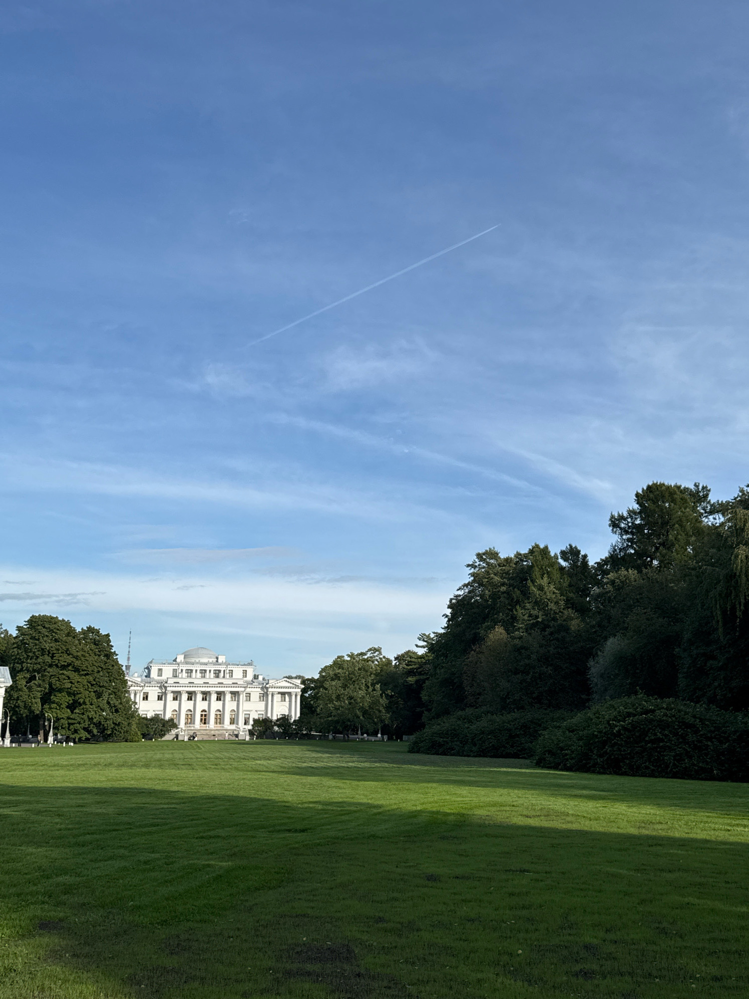 The image shows a wide, manicured green lawn in the foreground, with the sun casting shadows from the left. In the middle ground, a large, white neoclassical building with columns and a dome is visible, surrounded by lush green trees. A thin, white contrail from an airplane stretches across the bright blue sky. The building is identified as the Tauride Palace, located in Saint Petersburg, Russia. The scene depicts a serene park environment on a clear day.