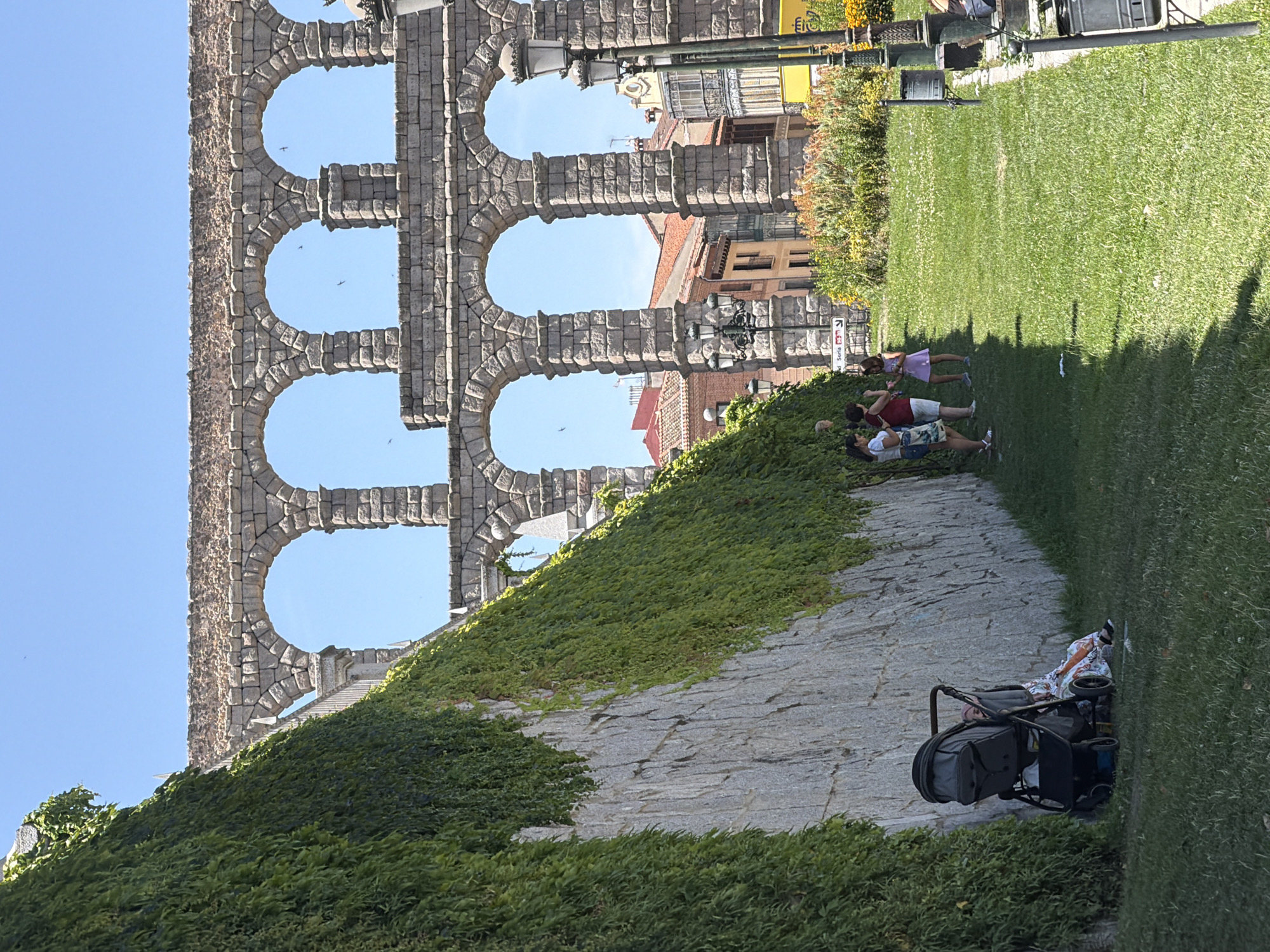The image captures a scene at the Aqueduct of Segovia, a Roman aqueduct located in Segovia, Spain. In the foreground, to the left, a large, grey stone wall covered in dense green ivy dominates the left side of the frame. A black stroller with a person seated next to it, dressed in a colorful dress, is positioned at the base of the wall. To the right, a lush green lawn slopes gently away from the wall. Three individuals, two adults and a child, are standing on the lawn near the wall, appearing to interact. Further in the background, the iconic aqueduct, constructed from massive granite blocks, stretches across the frame with its multiple tiers of arches. Beyond the aqueduct, the cityscape of Segovia is visible, featuring buildings with terracotta roofs. The sky is clear and blue. Lampposts and decorative flower beds are also present in the mid-ground and background.