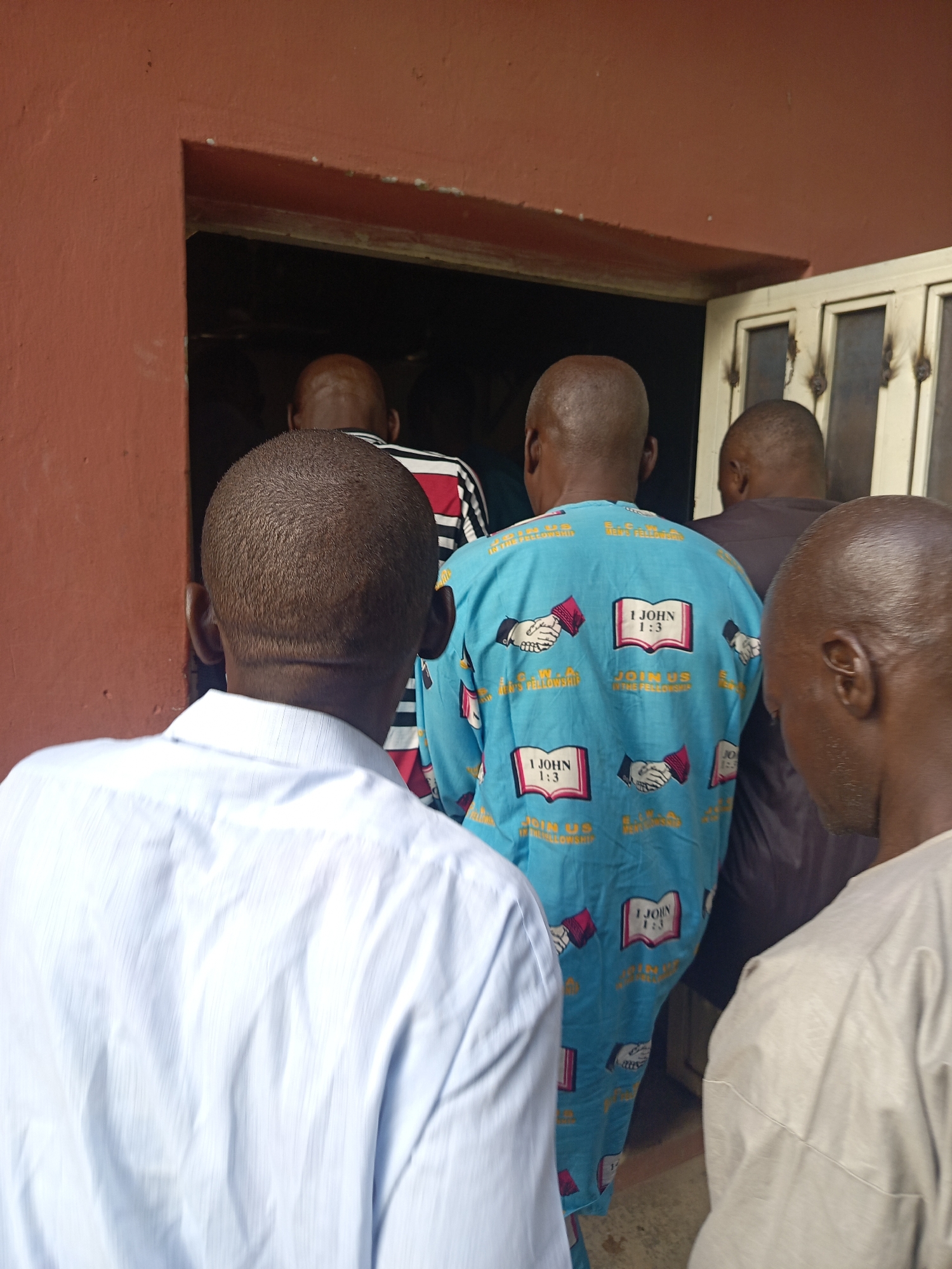 The image shows a group of men from behind, appearing to queue or enter a building in Akwanga, Nigeria. The scene is illuminated by bright natural light, indicating it is daytime. In the immediate foreground, a man with short, dark hair and prominent ears wears a light blue or white collared shirt. Directly behind him, another man is dressed in a vibrant, long-sleeved blue tunic. This tunic is adorned with repeating patterns that include open books displaying the scripture "1 JOHN 1:3," and an emblem featuring two clasped hands alongside the phrases "S.C.W.A. FELLOWSHIP" and "JOIN US IN THE FELLOWSHIP."
Further back, additional men are visible, including one wearing a black, red, and white striped shirt. The men are facing into a dark doorway, which serves as the entrance to a building. The exterior wall of the building is a distinct reddish-brown color, and a light-colored, possibly metallic, structure resembling a gate or window frame is partially visible to the right of the entrance. The collective posture of the men suggests they are entering the building for a communal event, likely a religious gathering or fellowship service, given the explicit biblical text and fellowship invitations on one man's attire.