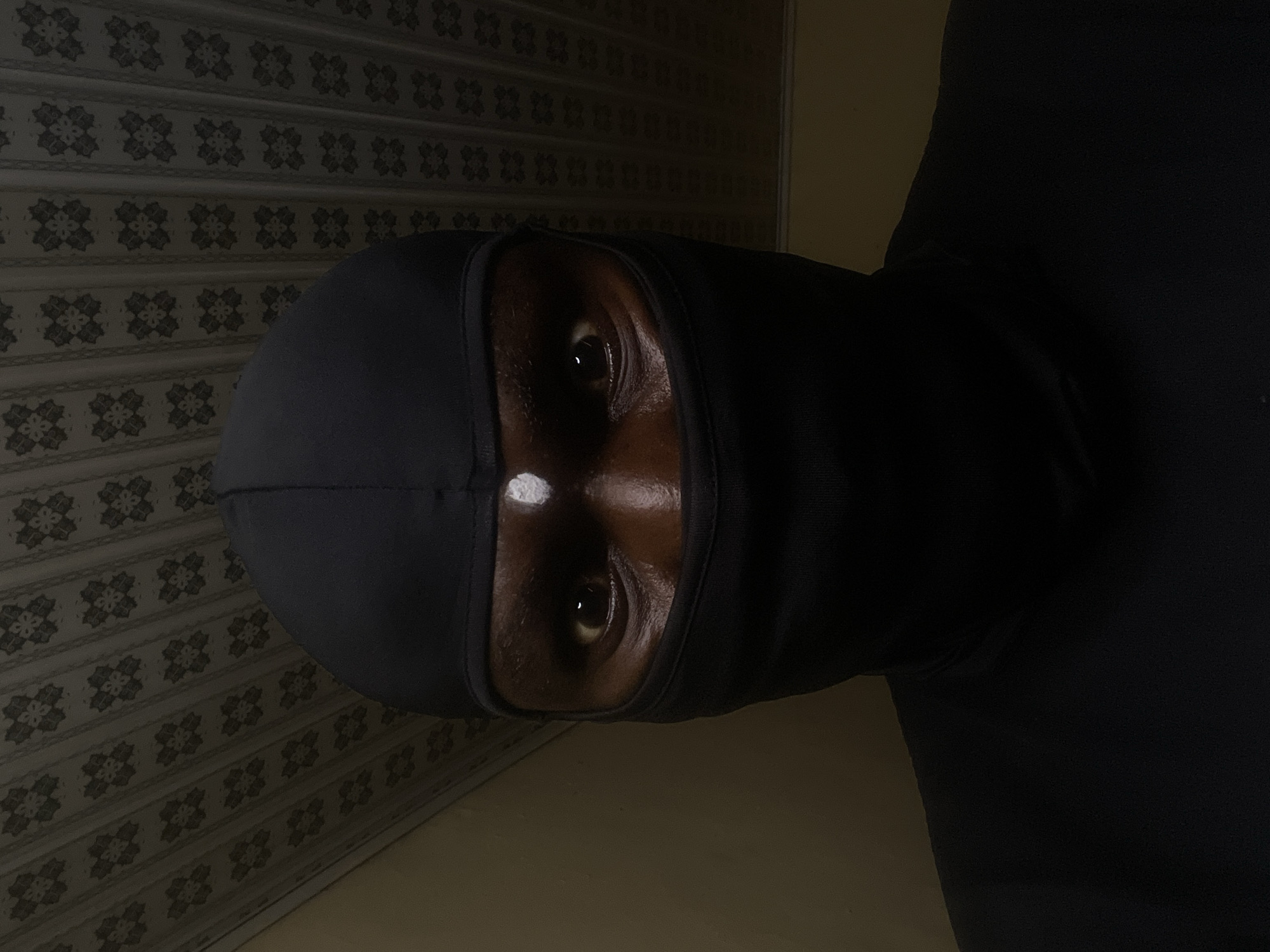 A person wearing a black balaclava is photographed indoors. The person's eyes and nose are visible through the mask opening. The person's skin is dark. There appears to be a small white mark or reflection on their nose. The background shows patterned wallpaper on a wall. A portion of a wall with lighter coloring is present in the lower right. No specific location information, such as the city of Jos, Nigeria, is discernable from the image itself.