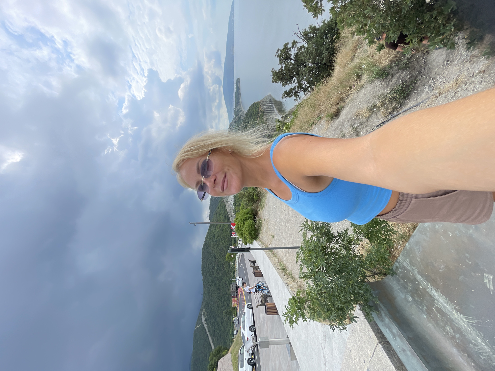 A blonde woman with sunglasses takes a selfie from a cliff overlooking the sea. She is wearing a blue tank top and brown shorts. In the background, a winding road with cars, a few benches, and a person on a bicycle are visible. The sea stretches out to the horizon, with a distant headland and a cloudy sky above. The location is the observation deck of the Mys Myshkako in Novorossiysk, Russia.