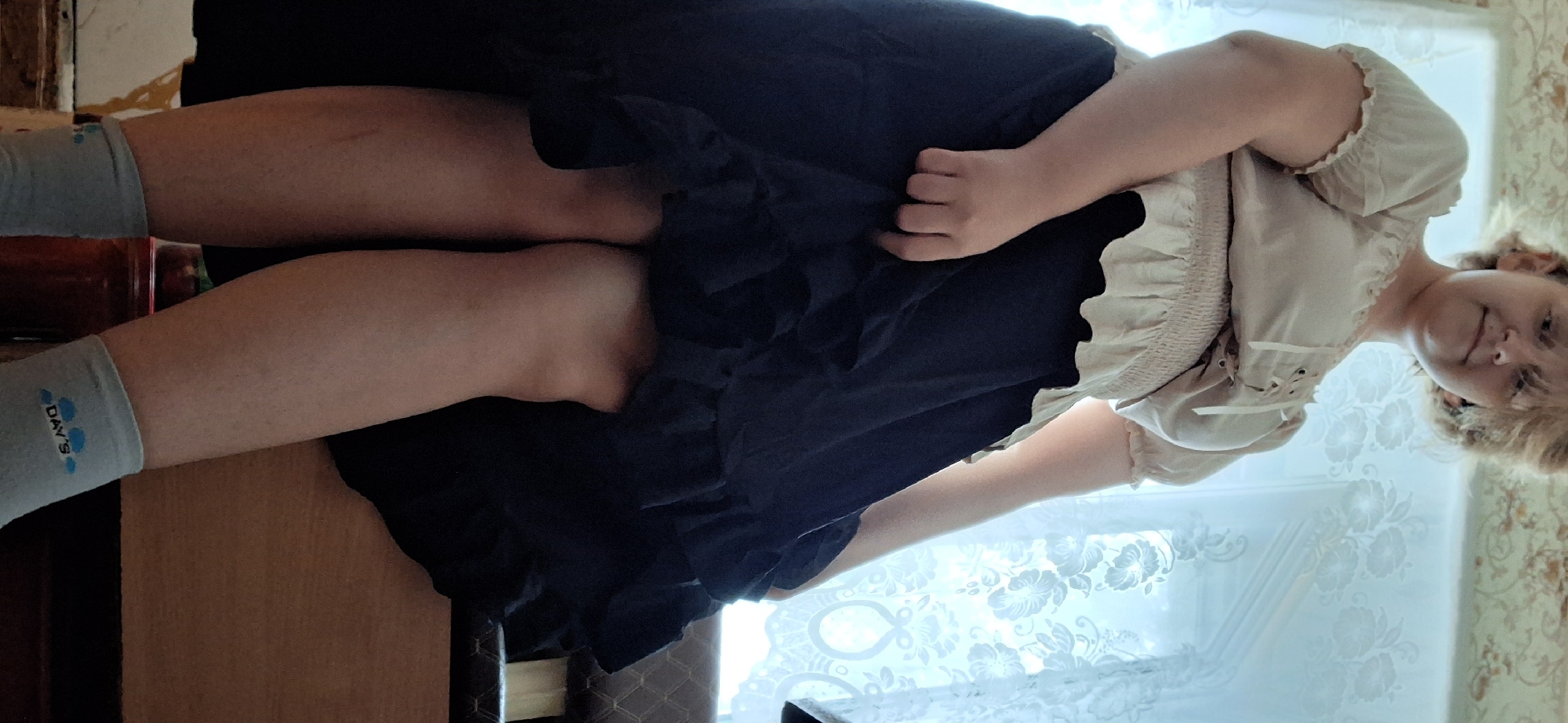 A person is shown from a low angle, looking up at them. They are wearing a light-colored, ruffled blouse with lacing at the front and a dark, ruffled skirt. Their legs are visible below the skirt, and they are wearing light blue socks with a cloud and "DAY'S" logo. The background includes a window with lace curtains and a patterned wallpaper. The scene appears to be indoors, possibly in a home in Petropavl, Kazakhstan.