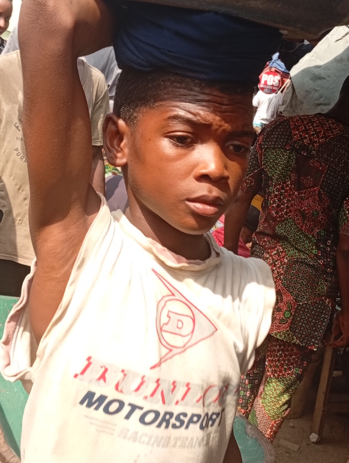 The scene is an outdoor, crowded setting in Onitsha, Nigeria. A young male, likely adolescent, is positioned in the foreground, facing slightly right with a neutral expression. He carries a dark, undefined object, possibly a basin, on his head, cushioned by a folded blue fabric. His right arm is partially raised. He wears an off-white sleeveless t-shirt printed with a red Dunlop logo and "MOTORSPORT RACING TEAM" text. To the immediate left background, a person in a tan t-shirt is partially visible. To the right, another individual wears a multi-patterned garment featuring green, red, and brown geometric motifs. Further in the right background, a person wearing a light-colored shirt has a red and white item marked "POS" visible on their back. The area is under direct sunlight.