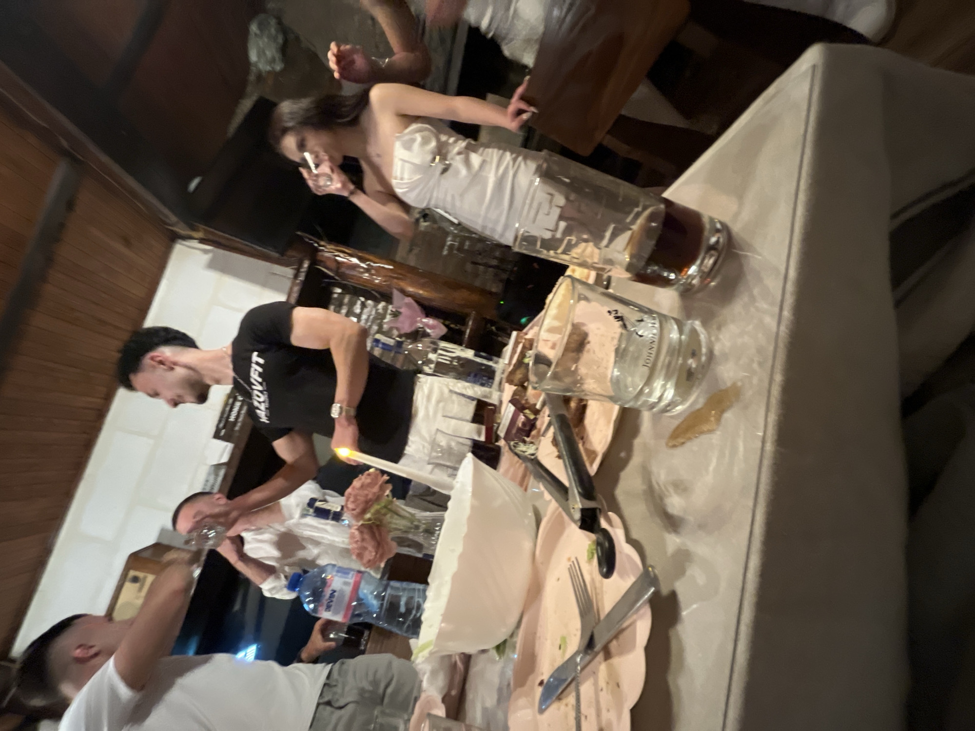 Indoor scene in Presoka, Bulgaria. Focus on a table laden with dishes, glasses, cutlery, a large bowl with remnants of salad, a bottle of Devin water, and flower arrangements with lit candles. Three men and one woman are visible. One man stands by the table wearing a black t-shirt. Another man, in a white shirt and gray pants, lifts a glass toward his mouth. A third man stands at the left with a white shirt and is holding a glass. The woman is wearing a white dress and holding a glass up to her mouth.