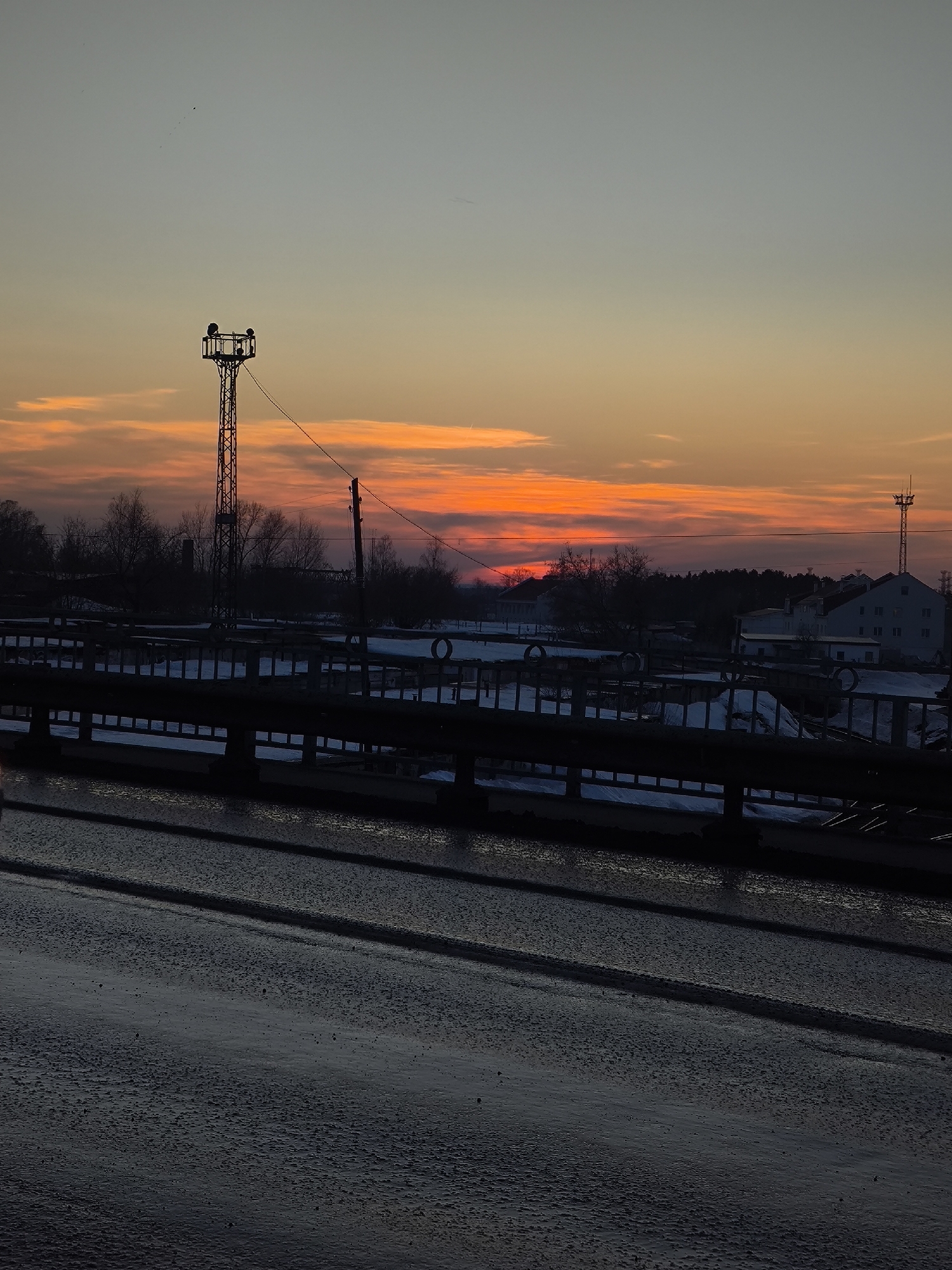 The image displays a winter twilight scene in Chaikovsky, Russia, characterized by a vivid sunset or sunrise. The foreground features a dark, wet asphalt surface, likely a road or bridge deck, showing visible texture and reflections. Three faint, parallel dark lines extend across this surface from the lower left to the right. Above this, a dark, horizontal railing or barrier runs across the lower middle of the frame.
Beyond the railing, the middle ground reveals a snow-covered landscape with various structures. On the left, a tall, latticed metal tower with an observation platform and attached equipment stands prominently. Further to the right, utility poles with overhead wires are visible. Bare trees, silhouetted against the bright sky, form a dark line along the horizon. Several low-rise buildings, including multi-story structures with light-colored facades, are discernible among the trees and snowdrifts on the right side. Round, dark objects, possibly tires or pipes, are partially embedded in the snow.
The sky in the background transitions from a pale blue-grey at the top to vibrant orange and deep red hues closer to the horizon, indicative of a sunset or sunrise. Horizontal cloud formations are illuminated in intense shades of orange and red, with the sun's direct light source appearing just below or obscured by the horizon line. No people, actions, or specific interactions are visible within the scene.