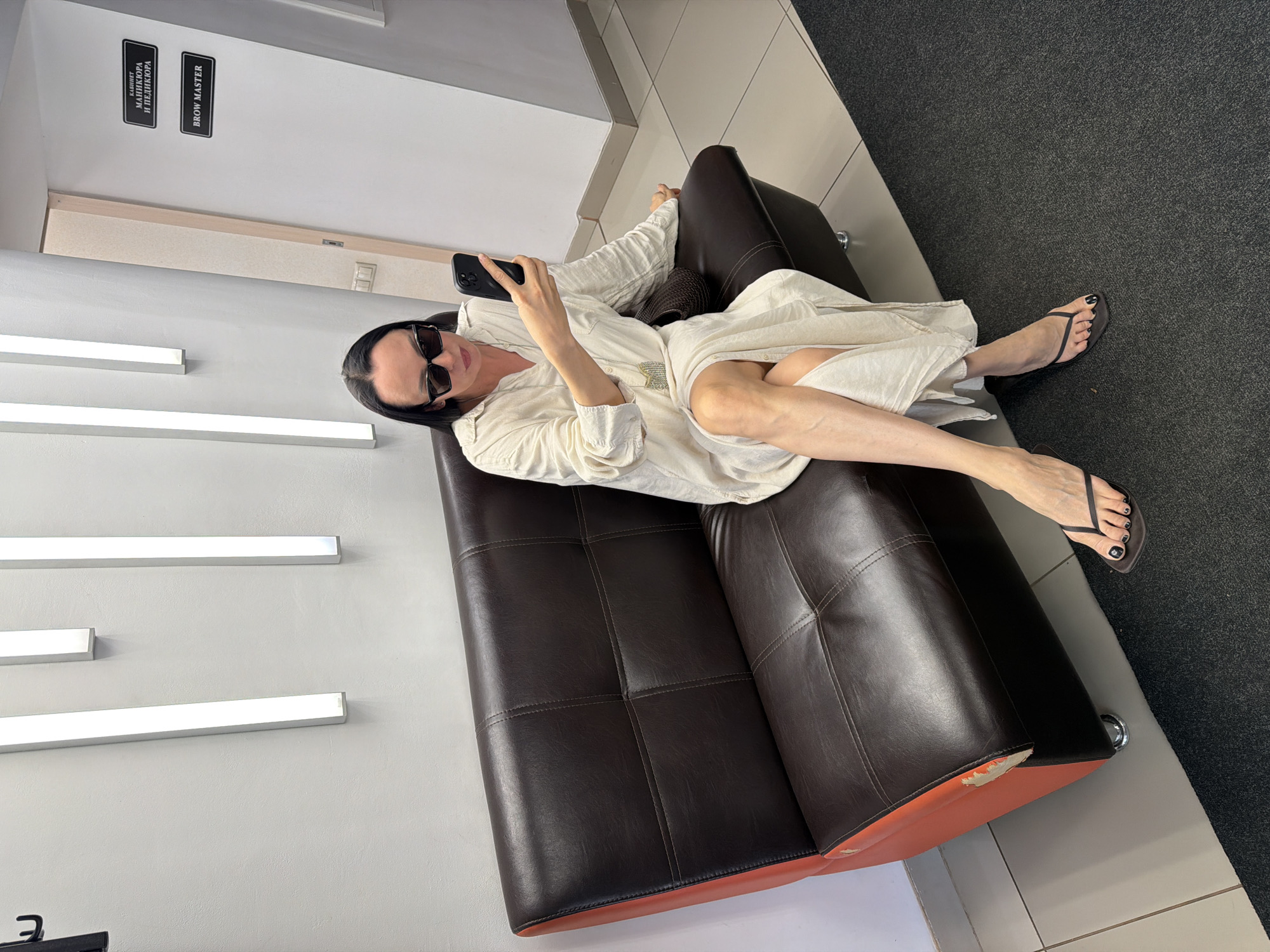The image shows a woman lounging on a dark brown leather couch in an indoor setting. She is wearing sunglasses, a light-colored loose shirt, and black flip-flops. She is holding a black smartphone in her hand. Her toenails are painted black. The couch has a tear at its lower corner, revealing an orange base. The room has white walls with horizontally aligned rectangular light fixtures. Two signs are present on the wall; one says "BROW MASTER" and the other one says "КАБИНЕТ МАНИКЮРА И ПЕДИКЮРА" in Russian. The floor is partly covered with grey carpet. The setting appears to be a beauty salon or similar establishment located in the city of Chaikovsky, Russia.