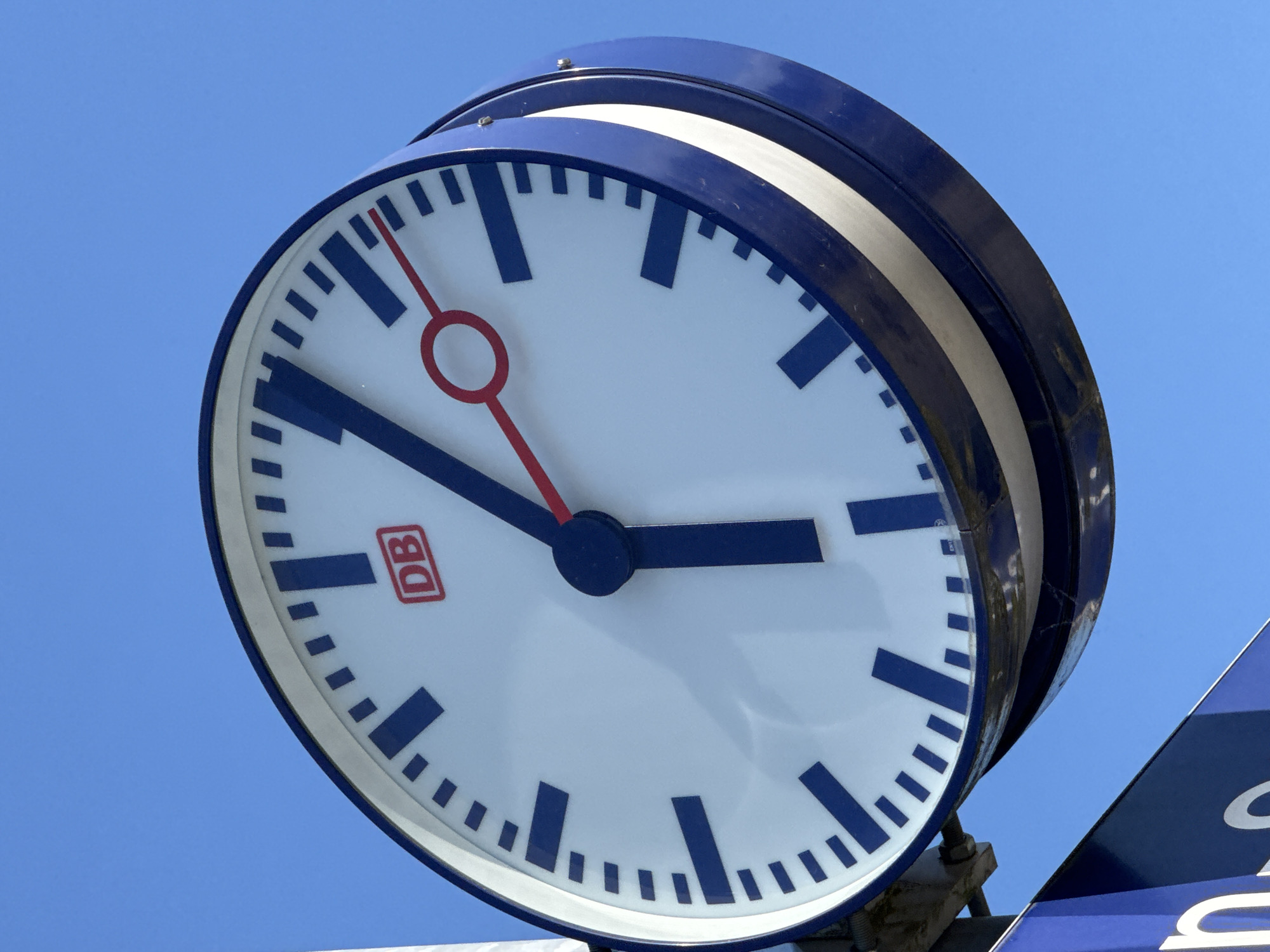 This media file is a close-up, angled shot of a large, round clock mounted outdoors against a clear blue sky. The clock has a white face with thick blue hour markers and smaller blue minute markers. The clock hands are dark blue, with a thinner red second hand. The clock has a prominent blue outer rim with a white stripe. The logo "DB" in a red square is visible on the clock face. The time indicated is approximately 2:38. The clear blue sky suggests it is daytime with fair weather. The "DB" logo is a strong indicator that the location is a train station in Germany, specifically likely belonging to Deutsche Bahn. The overall impression is that of a functional public clock, likely at a train station in Höhenkirchen-Siegertsbrunn, Germany.