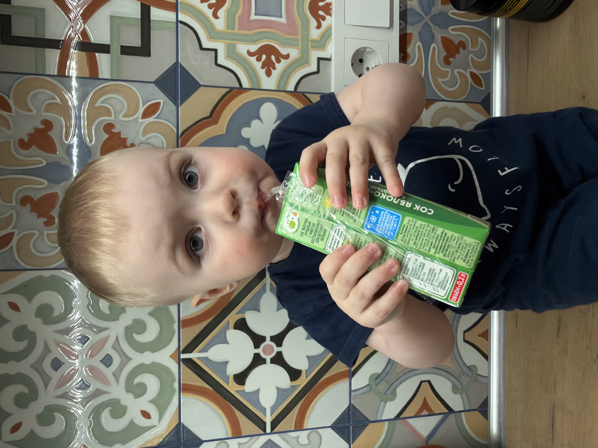 A young child with light blonde hair and blue-grey eyes is depicted in a close-up, top-down view, drinking from a green juice box through a straw. The child has rosy cheeks and is wearing a dark blue t-shirt with partial white text, which appears to spell "FLOW ALWAYS". Their hands are wrapped around the juice box, holding it steady as they sip.
The juice box is green and features Russian text, indicating it is "СОК ЯБЛОЧНО-ГРУШЕВЫЙ с мякотью" (Apple-Pear Juice with pulp), "ВОССТАНОВЛЕННЫЙ ДЛЯ ДЕТСКОГО ПИТАНИЯ" (Reconstituted for Baby Food), with a "ОБЪЁМ - 0,2 л" (Volume - 0.2 L). Other visible instructions include "ПОСЛЕ ВСКРЫТИЯ УПАКОВКИ ХРАНИТЬ НЕ БОЛЕЕ СУТОК ПРИ ТЕМПЕРАТУРЕ" (After opening, store for no more than 24 hours at temperature...).
The background features a tiled wall or floor with a vibrant, intricate pattern of geometric and floral designs in shades of blue, orange, green, and grey. A light-colored electrical outlet is visible on the tiled surface. To the right, a wooden surface, possibly a table or floor, and a metal edge are visible. The setting appears to be an indoor space, likely a kitchen or dining area in Votkinsk, Russia. The child looks directly at the camera with an intent, perhaps curious, expression. The lighting suggests it is daytime.