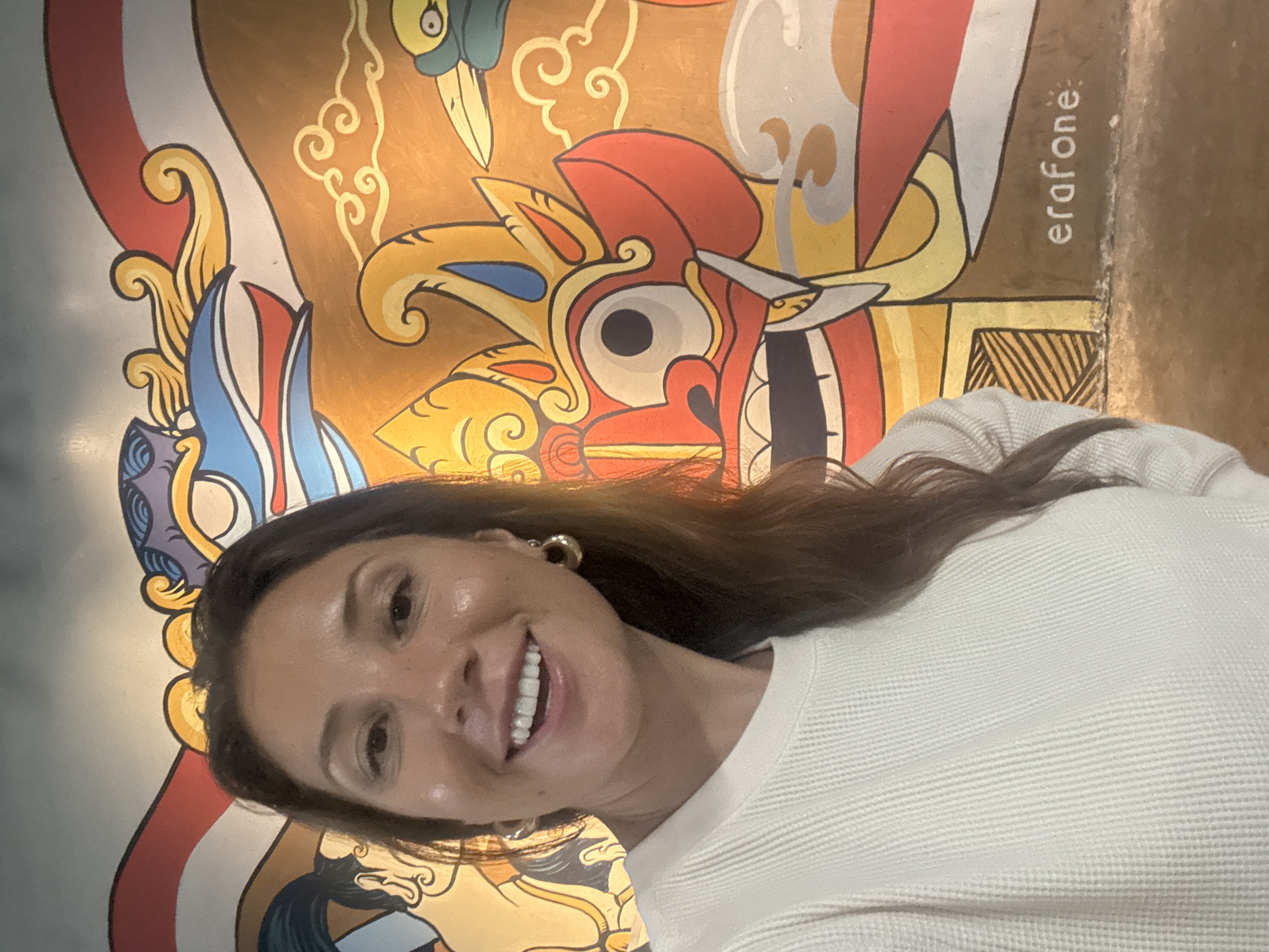 The media file is a portrait of a woman taken against a colorful mural. The woman, with long brown hair, is smiling and wearing a white waffle-knit shirt and large hoop earrings. She is positioned in the foreground, with her face slightly angled towards the camera. Behind her, the mural depicts a stylized depiction of a creature or mask, characterized by bold colors like red, yellow, blue, and white, with swirling patterns and intricate details. The mural has a golden background and appears to be painted on a wall. The text "erafone" is visible vertically on the right side of the mural. The scene suggests an indoor setting, possibly a cafe, restaurant, or art space, given the mural's presence. The lighting appears warm and even. The woman's expression is happy and relaxed, and the overall mood of the image is cheerful and vibrant due to the colorful background. The location context is Kuta Selatan, Indonesia.