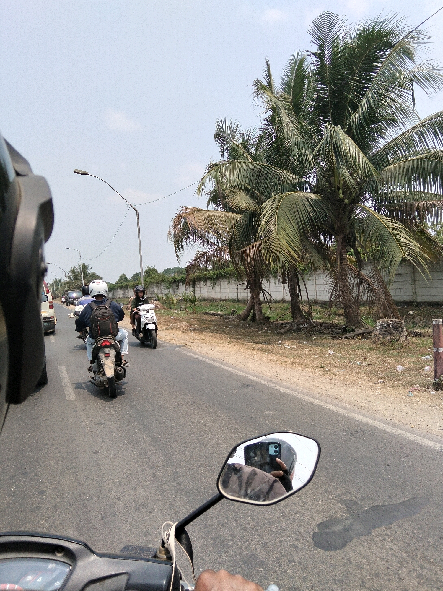 The image depicts a street scene in Sungai Rebo, Indonesia, characterized by a road with traffic and surrounding vegetation. In the foreground, the perspective is from the viewpoint of a motorcycle rider, with a partial view of the motorcycle's handlebars and a wing mirror. The wing mirror reflects the rider wearing a helmet and holding a smartphone, which is also capturing the scene.
The road is asphalt, with white lane markings. Several other motorcycles are visible on the road. One motorcycle, directly ahead of the viewpoint, carries a rider with a backpack and a white helmet. Another motorcycle is to the right, with a rider wearing a helmet and a dark shirt. A white car is present in the distance on the left side of the road.
The environment features tall coconut palm trees with their fronds reaching towards a partly cloudy sky. A low concrete wall runs along the side of the road, separating it from the vegetation. The ground beside the road is dry and covered with sparse grass, some debris, and a few plastic bottles. A street lamp is visible on the left side of the road. The overall impression is a daytime scene on a moderately busy road in a tropical or sub-tropical location.