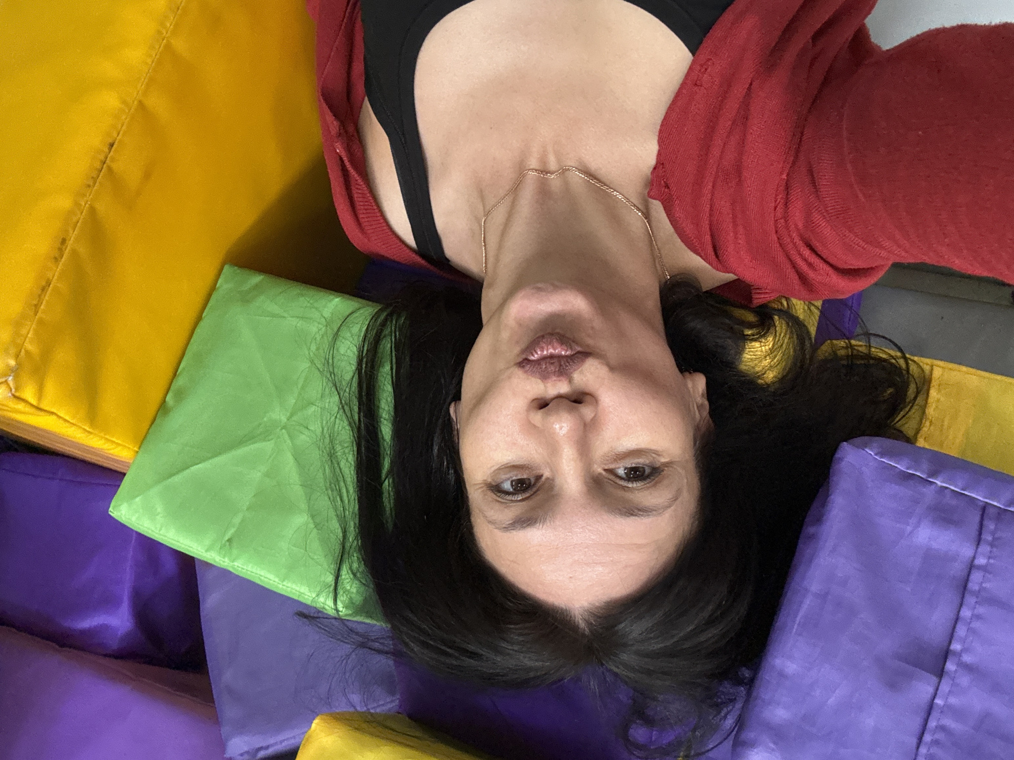 The media file is a close-up, upside-down selfie of a woman lying on a pile of colorful, soft rectangular cushions. The cushions are in vibrant shades of yellow, green, and purple, creating a playful and inviting visual. The woman has long, dark hair that is spread out around her head. She is wearing a red cardigan over a black top and a delicate gold chain necklace. Her expression is somewhat pouty, with her lips pursed. The lighting suggests it could be indoors, possibly in a studio, gym, or playroom given the nature of the cushions. There is no visible text. The setting appears to be Chaikovsky, Russia. The image conveys a sense of casualness and perhaps a moment of lightheartedness or relaxation.