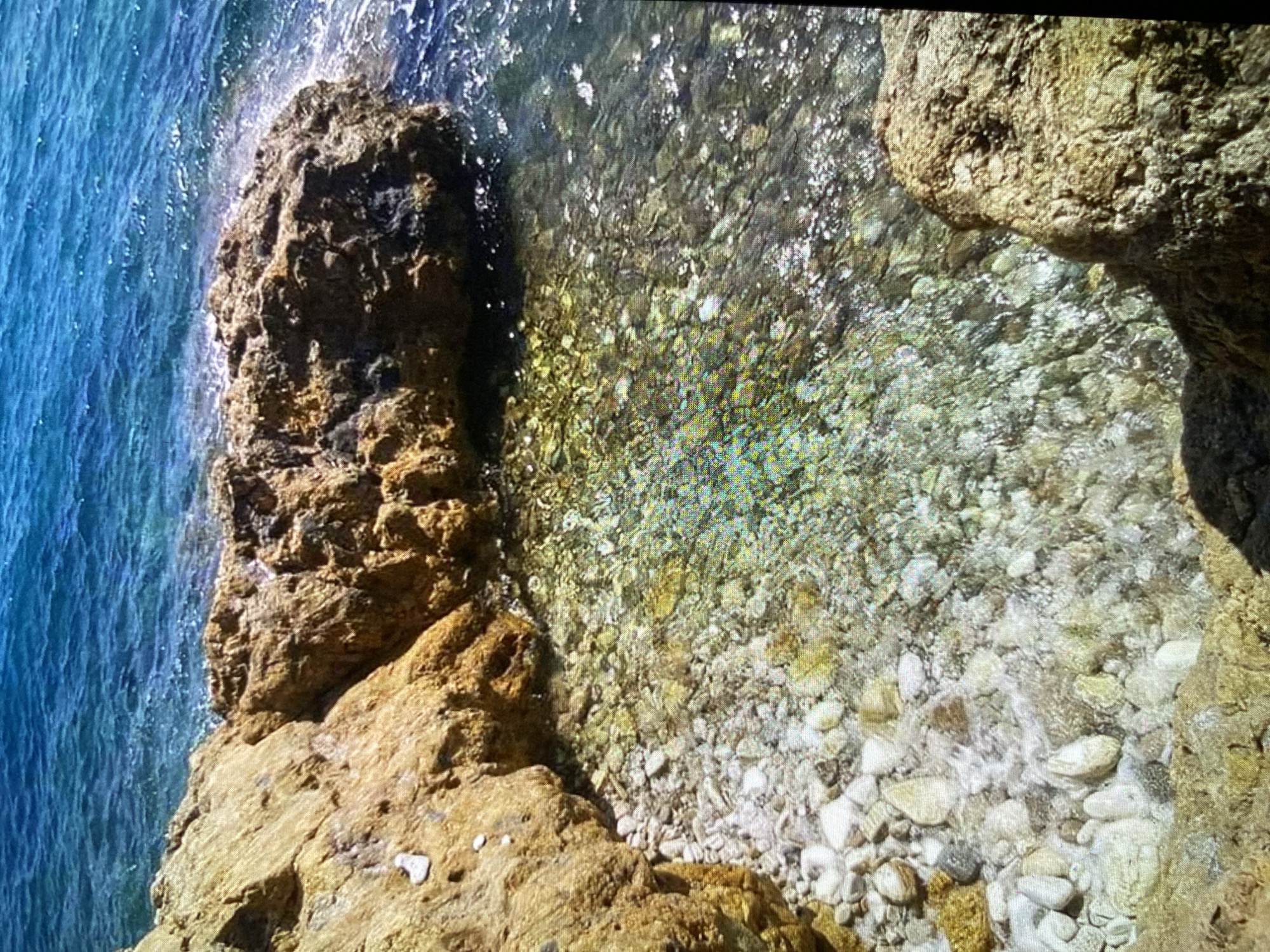 The image presents a vibrant, sunlit aerial view of a rocky coastline meeting clear, tranquil water. The scene is dominated by rugged rock formations and the translucent sea, revealing the seabed.
On the left side of the frame, the water transitions from a deeper blue further out to a lighter, clearer turquoise closer to the shore. The exceptional clarity of the water allows for a distinct view of the seabed, which is composed of countless pebbles and stones in various shades of white, grey, and light brown. Sunlight reflects brightly off the water's surface, creating shimmering patterns.
The right side and central area of the image are occupied by textured, weathered rock formations. These rocks are predominantly earthy brown and beige, with areas of darker grey and visible crevices, indicating their rugged nature. A large, prominent rock mass juts into the water, its base visibly interacting with the gentle lapping of small waves, which produce faint white foam. The underwater section of these rocks is also discernible through the clear water.
The lighting suggests a bright, sunny day, enhancing the natural colors and textures of the rocks and the water. There are no people, animals, or man-made objects visible in the image. No activity beyond the gentle movement of water against the rocks is apparent. There is no visible text. The overall impression is one of natural serenity and untouched coastal beauty.