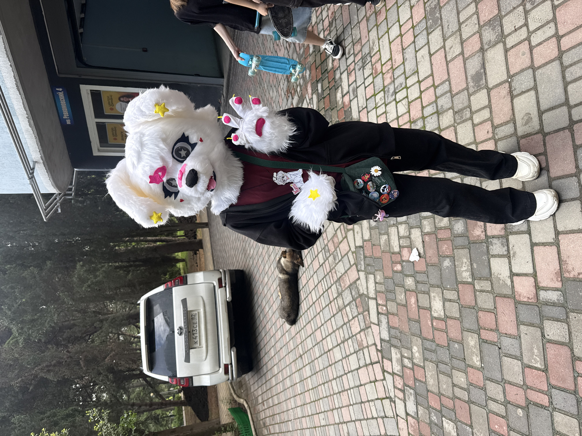 A full-bodied person dressed in a full-body fursuit stands at the edge of a brick-paved sidewalk, next to a parked white SUV with license plate number K413CE 82. The fursuit is white with a large head, decorated with pink and yellow star shapes, and large white gloves with pink fingertips. The person is wearing a black jacket, burgundy shirt, and black pants, as well as white sneakers. A green bag adorned with colorful buttons is slung across their body. A medium-sized dog lies still near the SUV's rear left tire. Another individual partially enters the frame, carrying a blue skateboard. The scene is set in an outdoor environment near a building with a sign bearing the inscription "ул. Революционный, 4". The background includes trees and greenery, suggesting a park or a similar outdoor space.