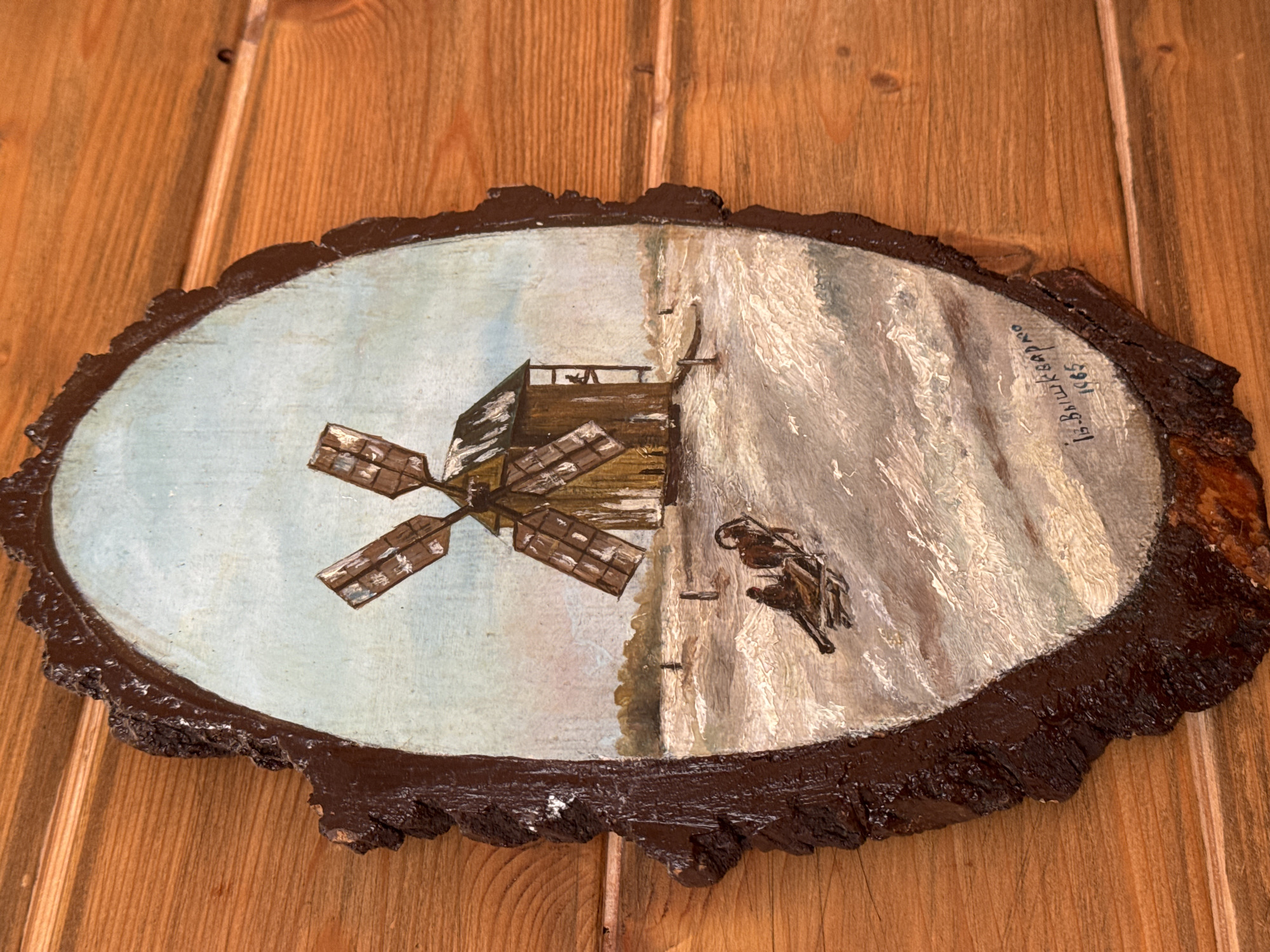 The image shows an oval painting on a slice of wood. The painting depicts a windmill on the left side and what appears to be a snowy mountain or cliff face on the right side. A figure, possibly on horseback or with a cart, is also painted near the cliff face. The painting is signed "I. Bishkerov 1965" in Russian. The wood slice has a bark border that has been painted dark brown. The artwork is mounted on a wooden plank wall. Ketoshi, Russia is not mentioned.