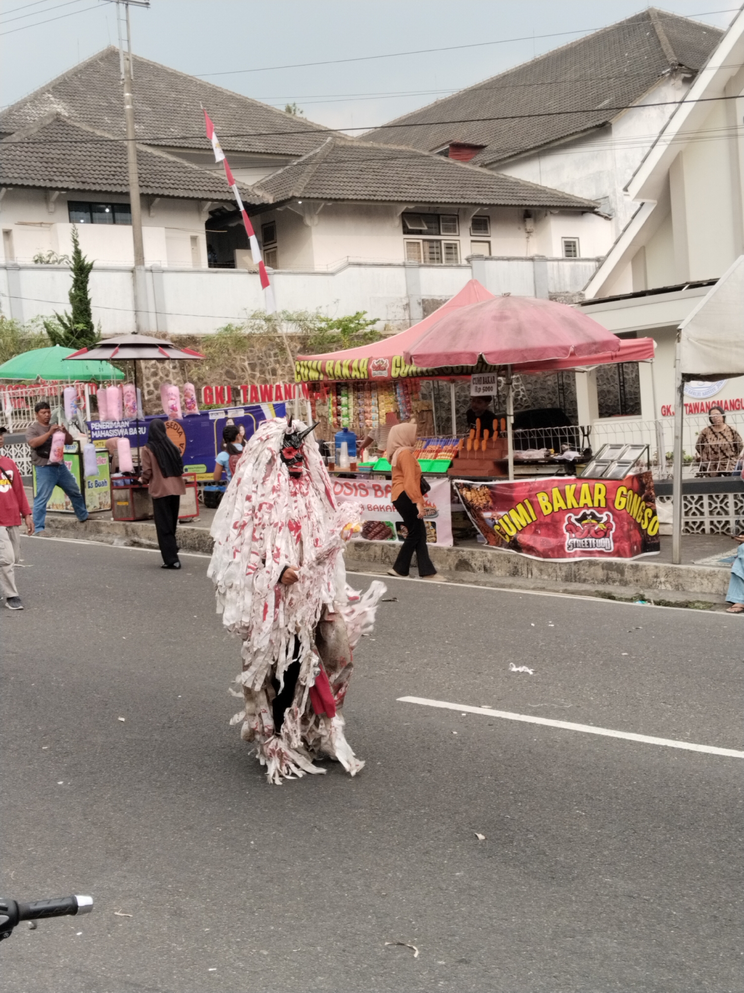 In Tawangmangu, Indonesia, a street scene unfolds with a costumed individual in the foreground, resembling a creature with a red and black mask and a flowing white costume adorned with red splashes, walking on an asphalt road. The person is positioned in the middle of the road, facing the viewer. Behind this figure, several food stalls are set up along the sidewalk, covered by red and green umbrellas. One stall displays a banner with the text "CUMI BAKAR GONGSO" and a cartoon logo of a squid. People are visible at the stalls, some interacting with vendors, and others browsing. In the background, a white building with a textured facade and a tiled roof is visible. A red and white striped pole with a flag is also present. The sky is overcast. The environment suggests an outdoor market or festival.