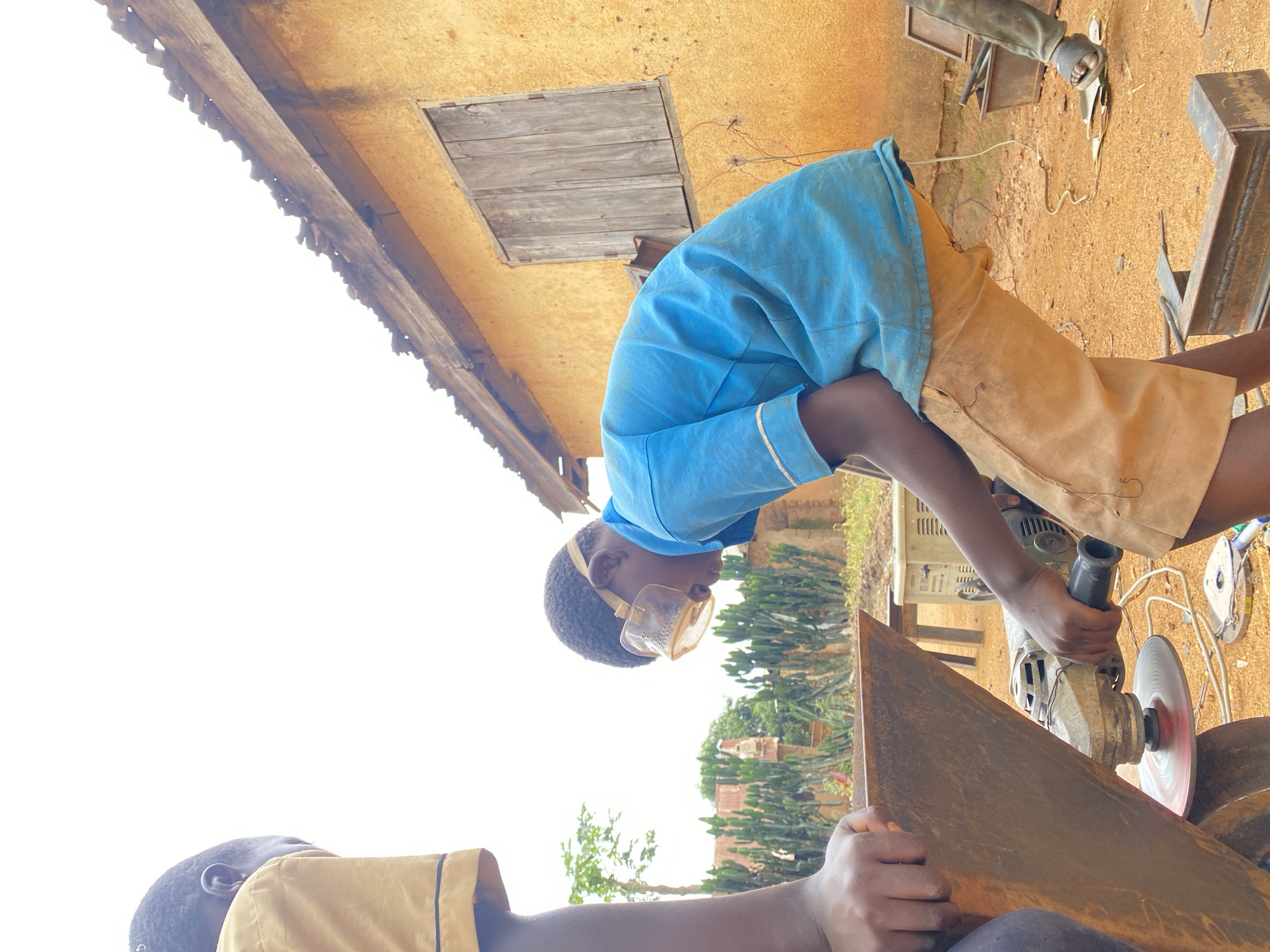 The image depicts an outdoor metalworking scene in Kuru Dungus, Nigeria, captured during daylight hours under a bright, possibly sunny sky. Two individuals, likely young, are engaged in cutting or grinding metal.
In the foreground and center, one person wearing a blue short-sleeved shirt, light brown or khaki shorts, and clear safety goggles operates an angle grinder. Their attention is focused downwards on a large, rusty metal sheet, which the grinder's disc is actively working on. The other person, partially visible in the bottom left, is wearing a light brown or khaki shirt and appears to be holding the metal sheet being worked on, or is closely observing the process.
The setting is characterized by a dusty, sandy ground. In the background, a building with a textured, light brown or yellow wall features a small wooden shuttered window. Scattered around the work area are various metal pieces, including what appear to be welded frames, electrical wires, and a box-like piece of equipment (possibly an old inverter or power supply). Further in the background, sparse green vegetation, including some cacti-like plants and a small tree, is visible beneath an overexposed white sky.
The scene conveys a sense of practical work or skill-building, with safety precautions evident in the form of the goggles worn by the grinder operator. There is no discernible text visible in the image.