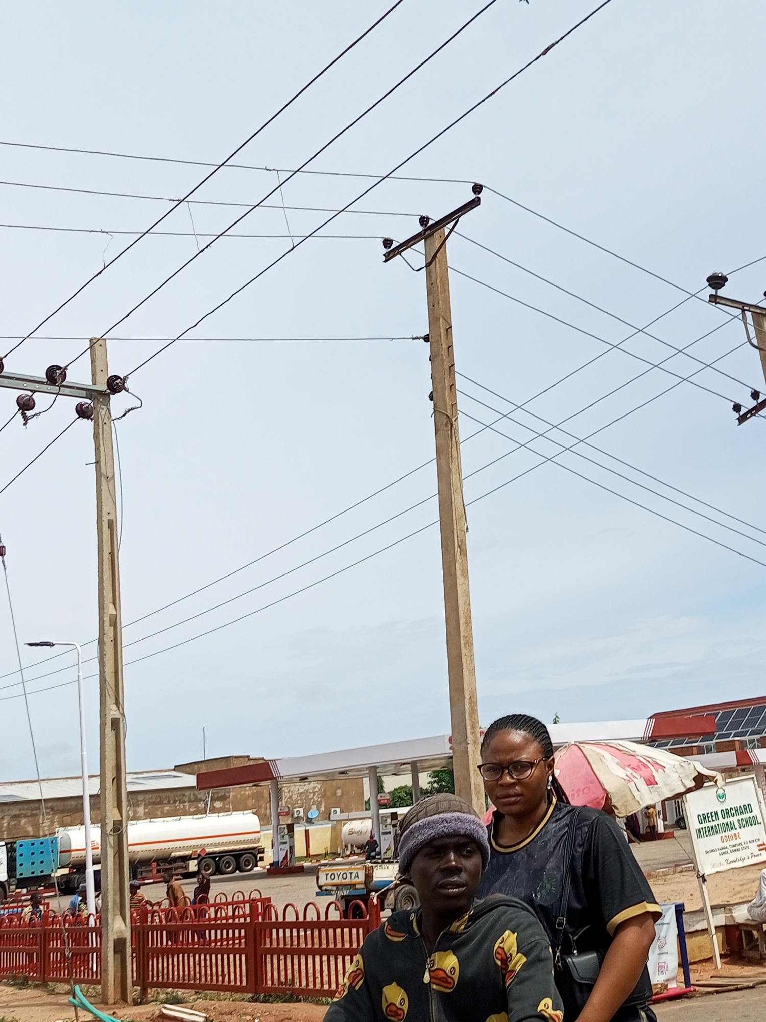 This image depicts a street scene in Tempure, Nigeria, likely near a gas station. In the foreground, a man wearing a patterned hoodie and a beanie is seated, looking to his left. To his right, a woman with glasses and dark clothing stands, looking forward. Behind them, a red metal fence encloses an area with several people visible. Further in the background, a gas station with multiple pumps is operational, with tanker trucks parked at the pumps. Overhead, power lines crisscross the sky, supported by concrete utility poles. A sign for "GREEN ORCHARD INTERNATIONAL SCHOOL" is visible on the right side of the image. The sky is overcast.