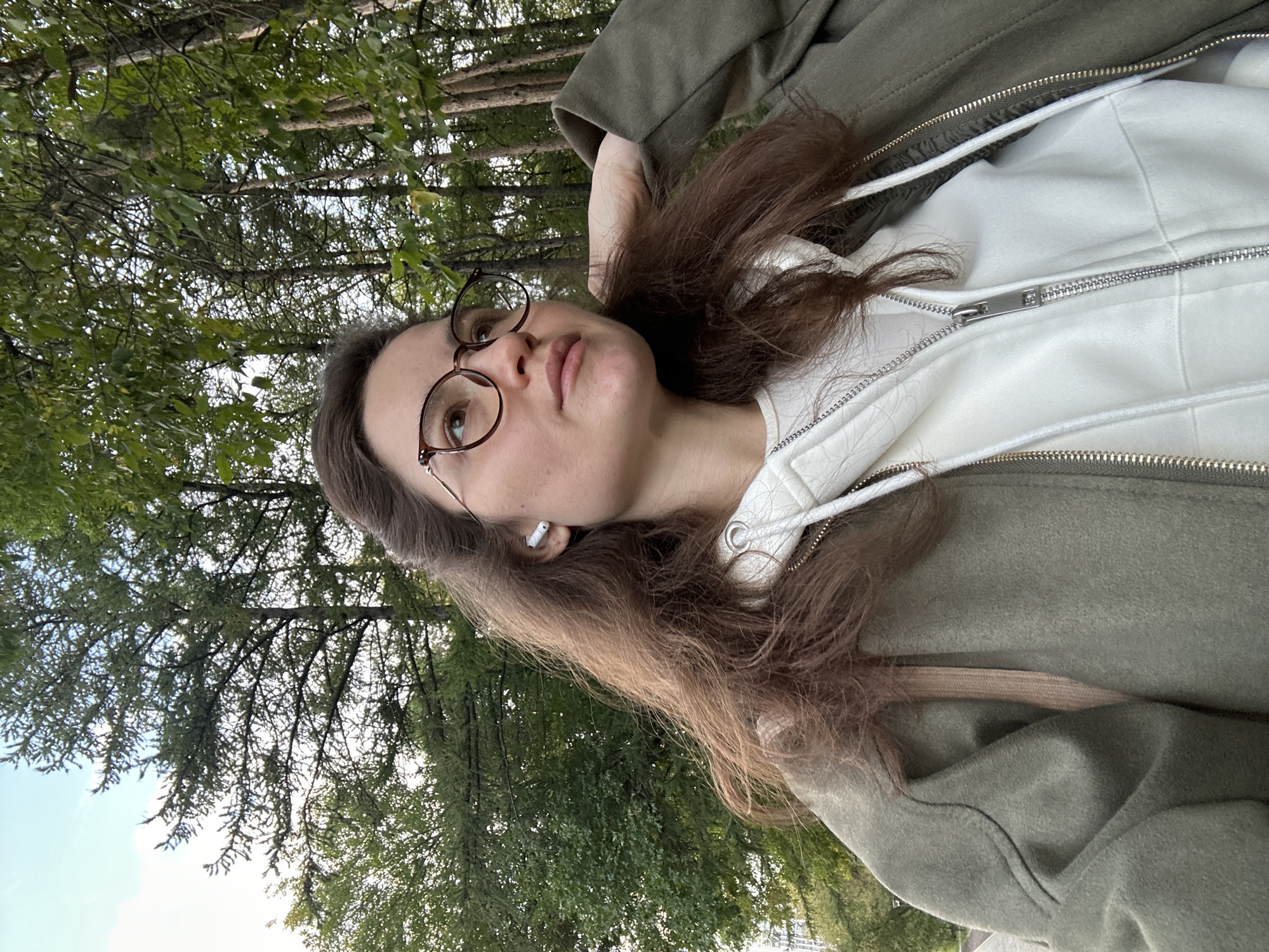 In an outdoor setting with trees and a cloudy sky, a young woman with long, dark brown hair and glasses is pictured. She is wearing a white hooded sweatshirt with a zipper and an olive green jacket. She has an earbud in her right ear and her right hand is resting on her hair. The scene appears to be in a park or wooded area, likely within the city of Ufa, Russia, given the context.