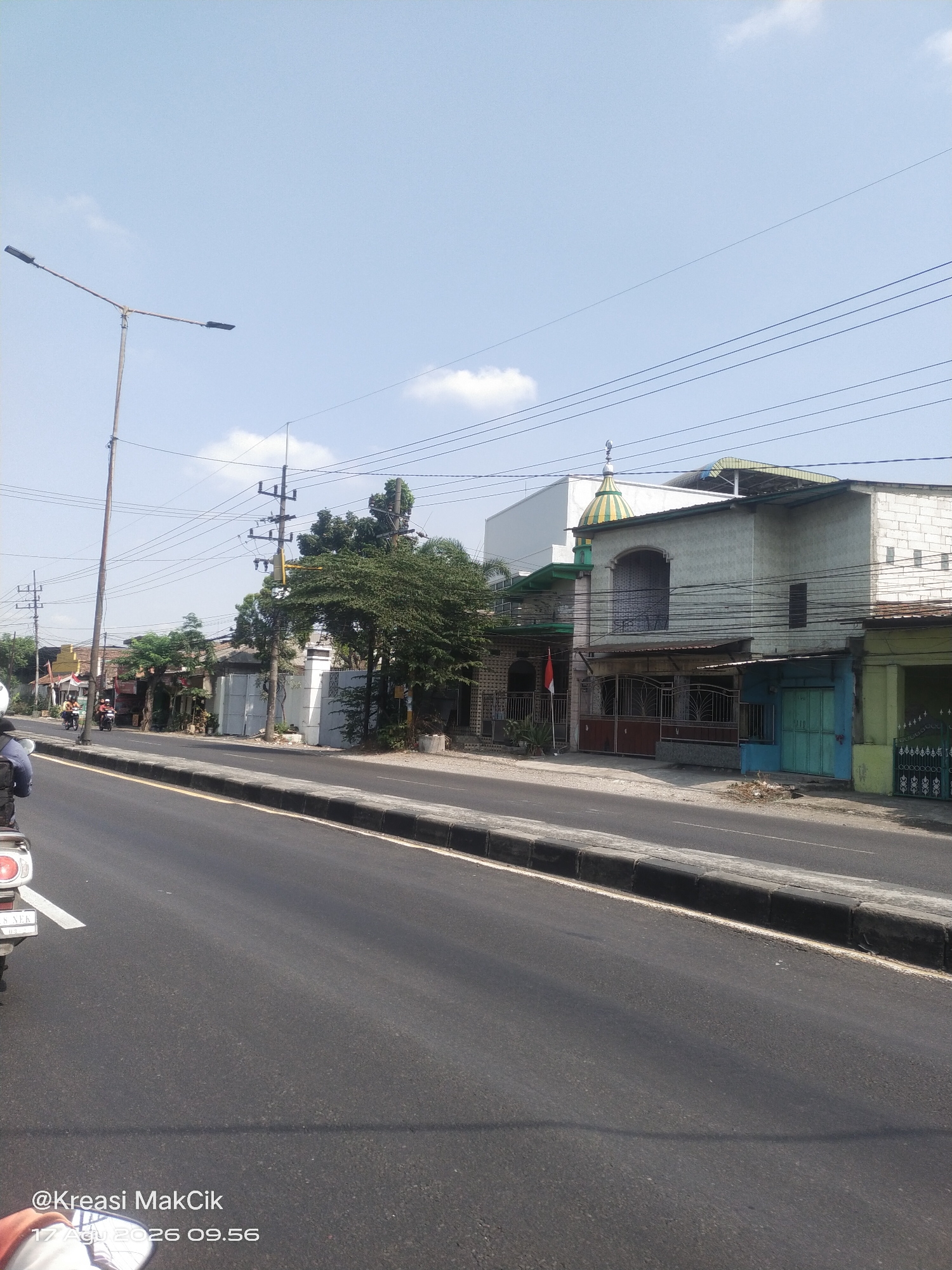 The image shows a street scene in Gilang, Indonesia. The foreground features a paved road with a concrete curb. On the right side of the road, there are several buildings with a mix of architectural styles, including some with tiled roofs and one with a green and yellow dome, possibly a mosque. There are also power lines overhead. On the left side of the road, a lamppost stands tall. Further down the road, several motorcycles are visible, with riders on them, suggesting ongoing traffic. The sky is clear blue with a few scattered clouds. The overall impression is a typical street view in a residential or mixed-use area of an Indonesian town on a sunny day.