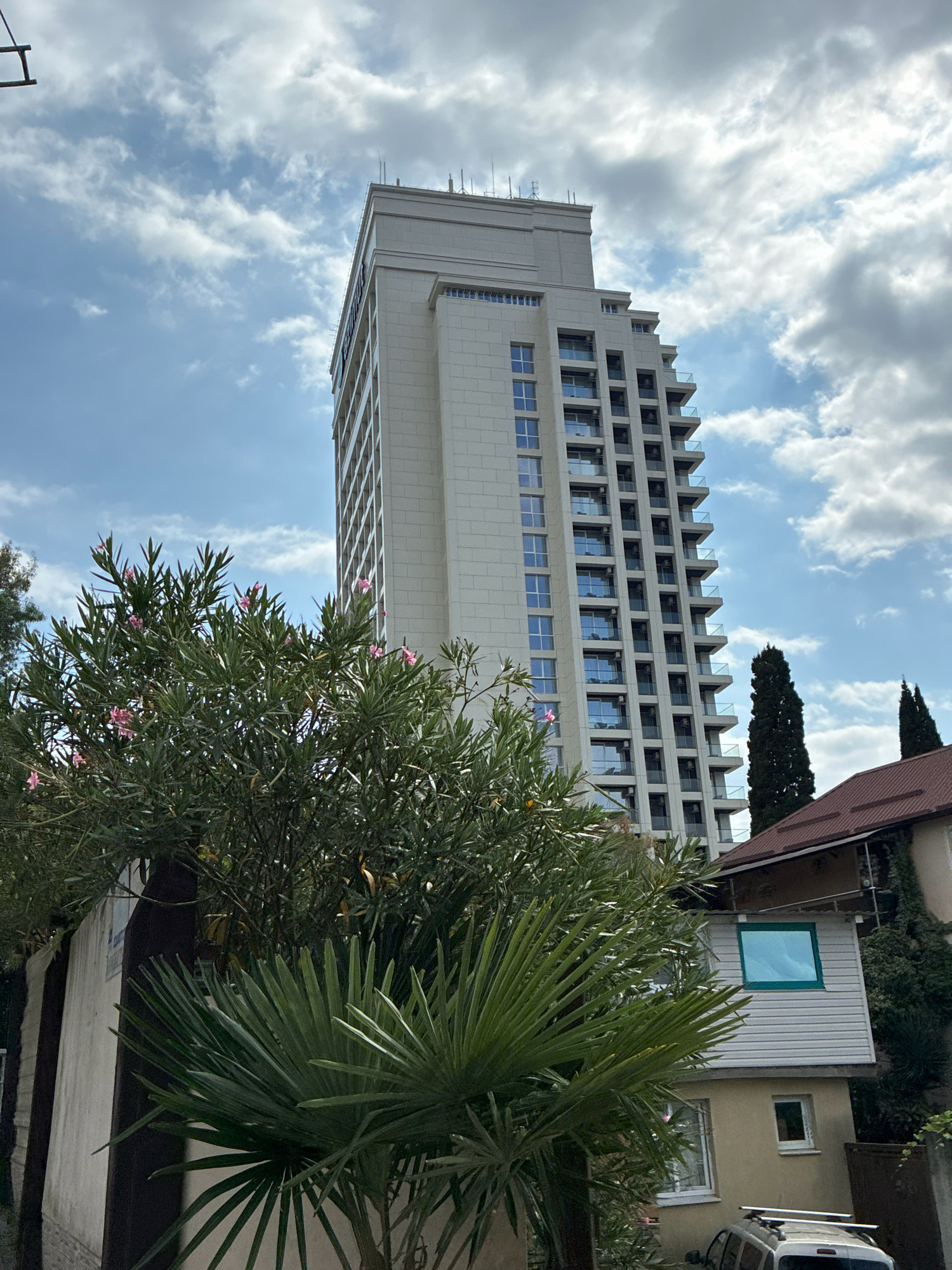 A tall, light-colored, modern apartment building with numerous balconies and windows dominates the upper portion of the image. It is set against a partly cloudy sky with scattered white clouds. In the foreground, lush green foliage from trees and shrubs partially obscures the view of the building and a smaller, older, tan-colored house with a brown-tiled roof. The house has a small green-trimmed window on an upper story addition and a smaller white-trimmed window on the lower level. A white car with a roof rack is parked in front of the house. The overall impression is a residential area with a mix of new and older construction, likely in a climate with abundant vegetation.