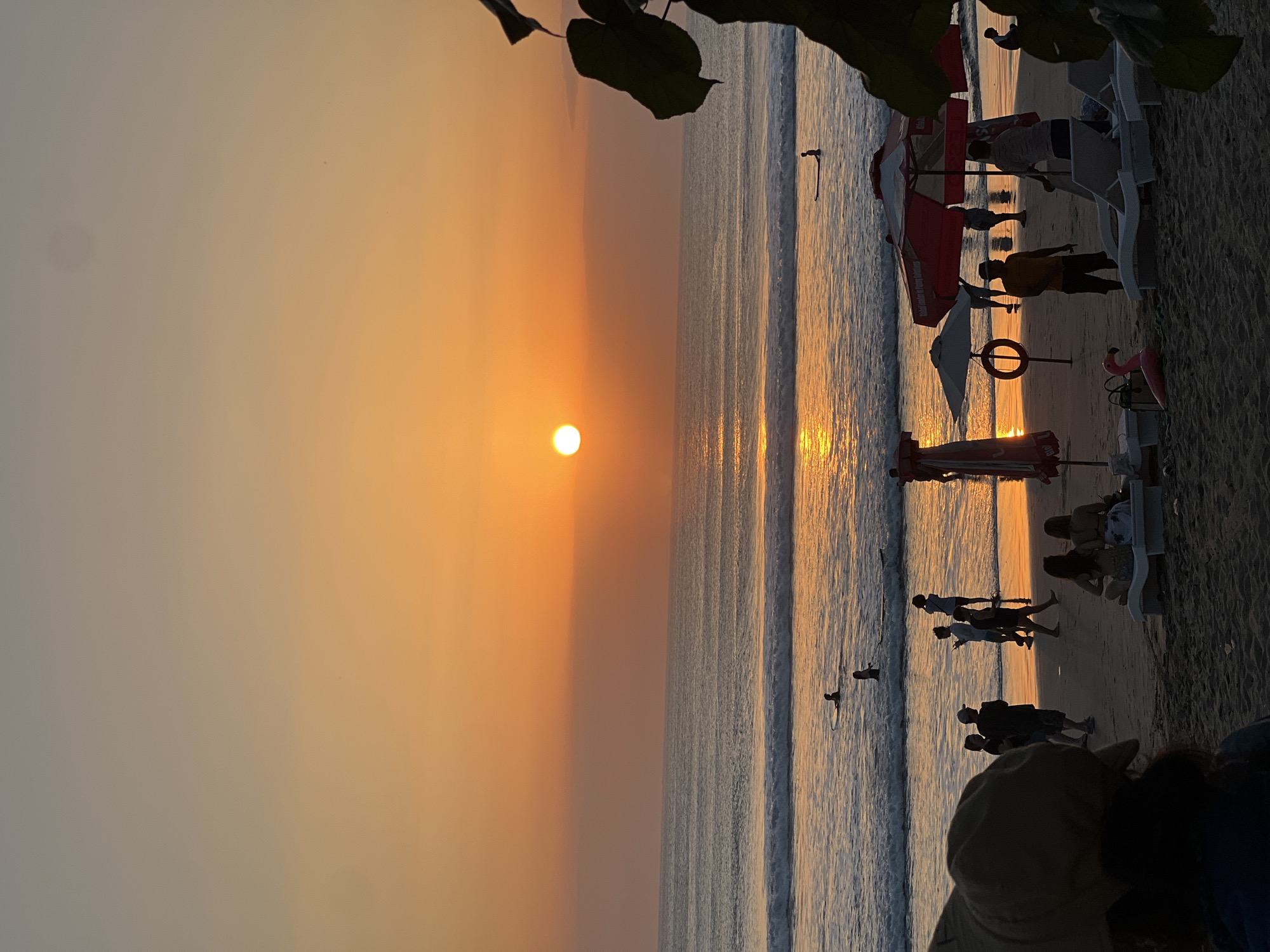 This image depicts a sunset scene at Kuta Beach in Kuta, Indonesia. The sun, a bright orange orb, is positioned in the center of the horizon, casting a warm glow across the sky and the ocean. The sky transitions from a pale orange near the horizon to a lighter hue of orange and then to a muted gray towards the top of the frame. Gentle waves are rolling onto the sandy shore, reflecting the sunset's light.
Several individuals are visible on the beach and in the water. In the water, a few people are seen with surfboards, suggesting surfing activities. Closer to the shore, a lifeguard stand with a red umbrella is present, and a red life preserver is mounted on a pole. Beach chairs and umbrellas are set up on the sand, some occupied by people. Silhouettes of people are scattered across the beach, some walking, some standing, and some sitting. A pink inflatable flamingo is also visible on the sand. Foliage from a tree is seen in the foreground on the right side, partially framing the scene. The overall atmosphere is one of a tranquil beach evening with people enjoying the sunset.