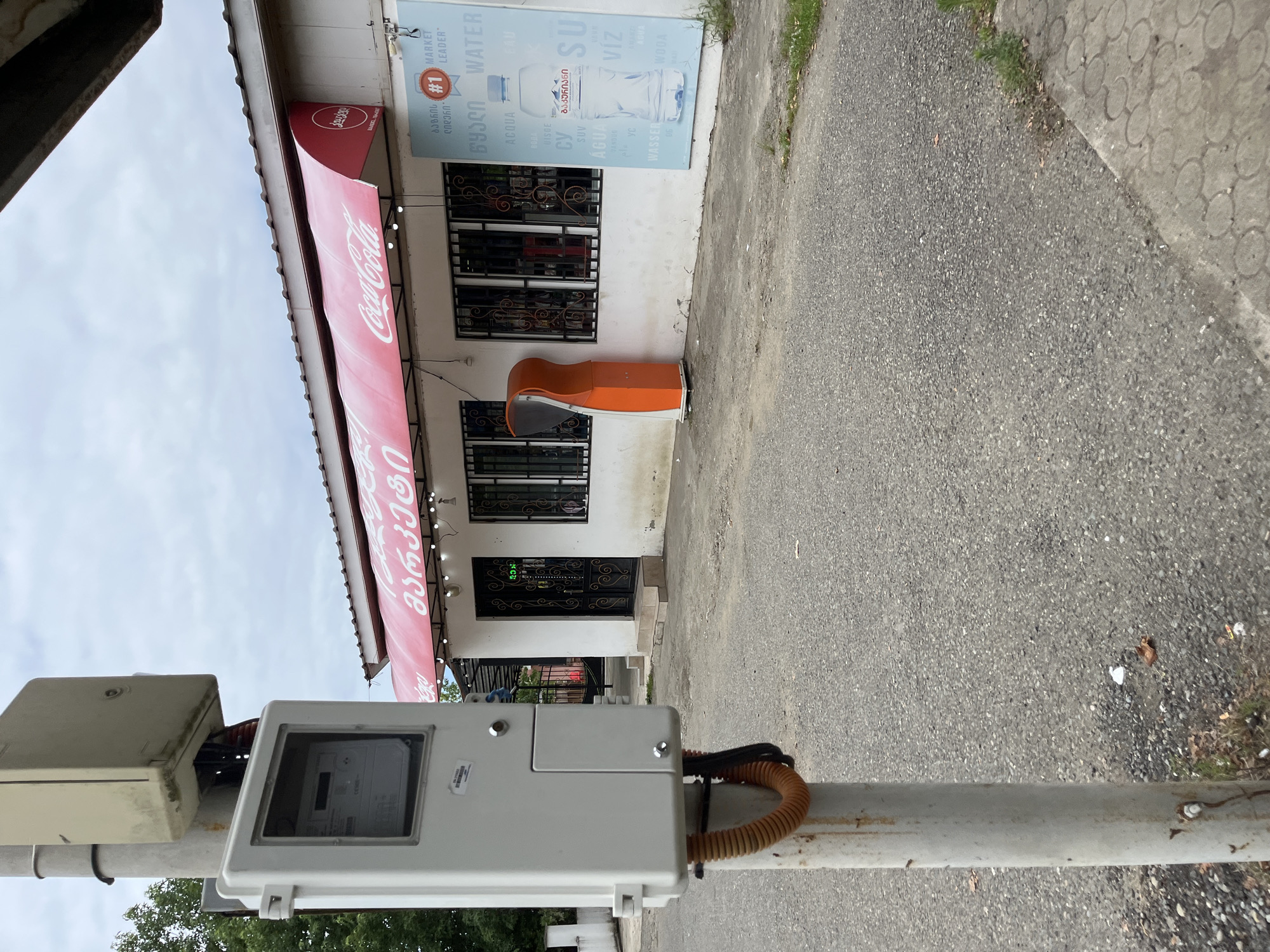The image shows a street scene in Ureki, Georgia, with a prominent outdoor electrical meter box on the left side of the frame. The meter box is mounted on a metal pole and has a gray casing with a clear window revealing the meter inside. Orange and black corrugated tubing connects to the bottom of the meter box. In the background, a white building with a red awning displays a "Coca-Cola" logo. A bright orange public telephone booth is situated to the right of the building's entrance. A large poster advertising water is affixed to the building's exterior, featuring a bottle of water and text in various languages, including Georgian. The ground in front of the building is paved with asphalt and a section of concrete sidewalk is visible on the right. Overcast skies suggest a cloudy day.