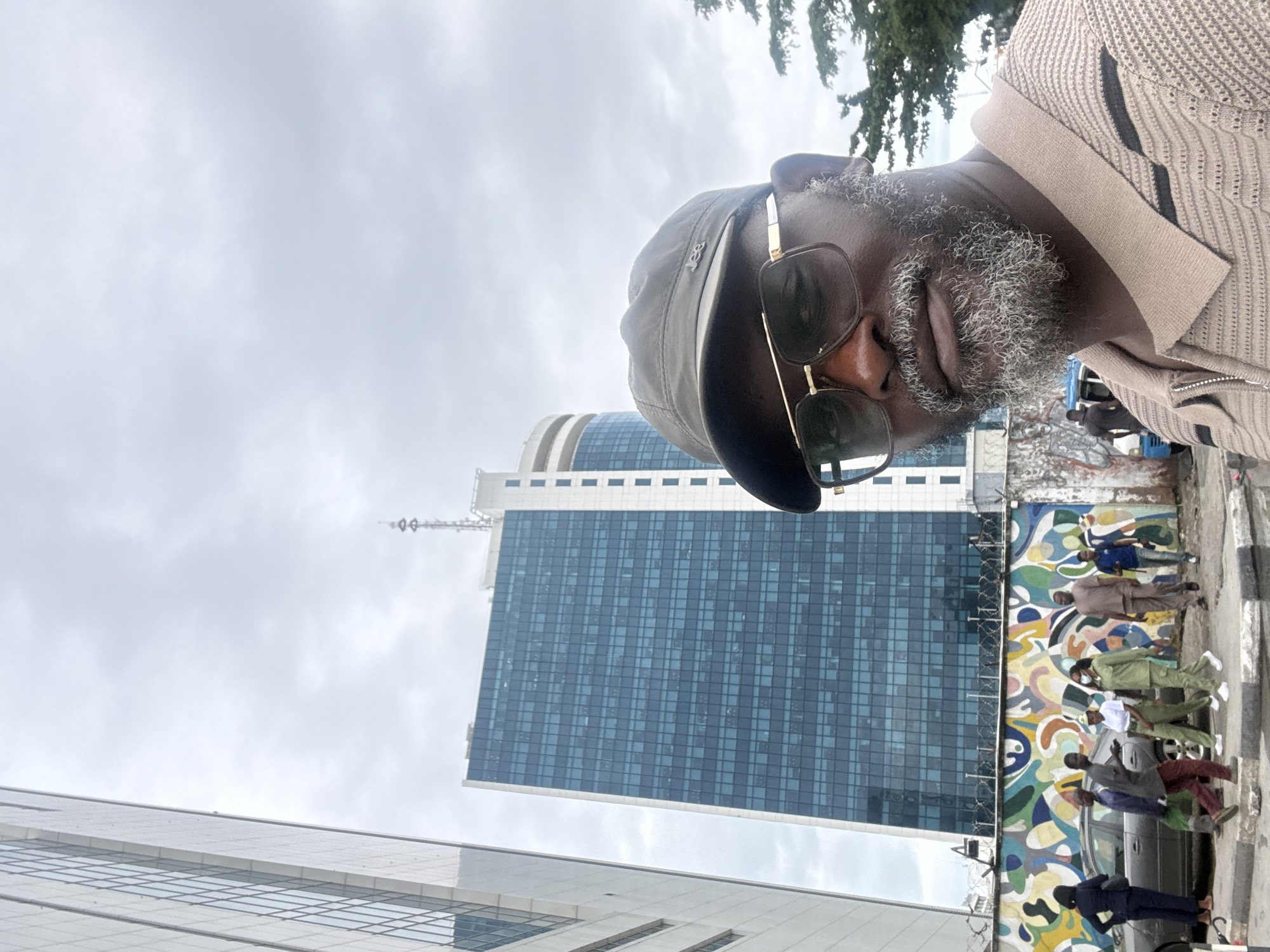 The media file is an image taken outdoors in Itirin, Nigeria, on a cloudy day. The main subject is a man looking towards the camera, with his head tilted. He is wearing sunglasses, a dark baseball cap, and a beige and black patterned sweater. He has a grey beard and appears to be middle-aged.
In the background, there are several buildings. A tall, modern skyscraper with a blue glass facade dominates the center of the image. To the left, another building with a glass exterior is partially visible. Beneath the skyscraper, there is a vibrantly painted mural on a wall. A group of people, some in uniform, are standing and interacting in front of the mural and near a vehicle.
The scene suggests a public space, possibly a street or plaza in an urban area. The overcast sky indicates it is likely daytime, but the specific time is not discernible. The overall mood is neutral, with no strong emotional cues evident from the subjects. No visible text is present in the image.