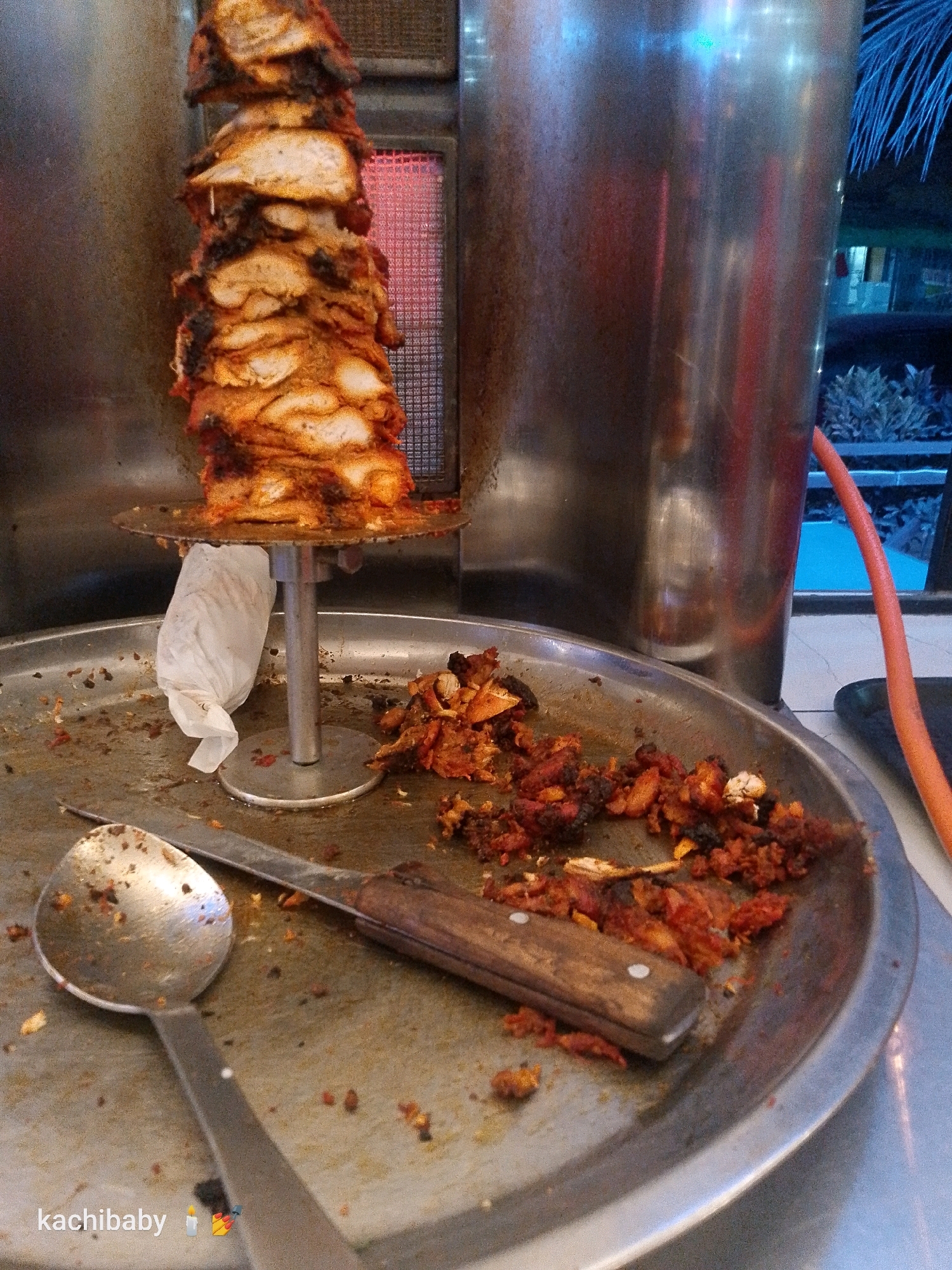 A close-up, eye-level shot shows a vertical rotisserie with seasoned chicken meat being cooked. Below the rotisserie, a large, circular metal tray catches drippings and scraped meat. A silver spoon and a knife with a wooden handle lie on the tray, alongside discarded pieces of cooked chicken. A white paper wrapper is attached to the rotisserie stand. In the background, a large metal cylinder, likely part of the cooking apparatus, is visible. To the right, an orange hose snakes out of frame. The environment appears to be an outdoor food stall in Shanzu ward, Kenya, as indicated by the "kachibaby" watermark and candle emojis in the bottom left corner of the image. The lighting suggests it may be evening or nighttime, with artificial lights illuminating the scene and some ambient light from what appears to be a dimly lit background with palm fronds.