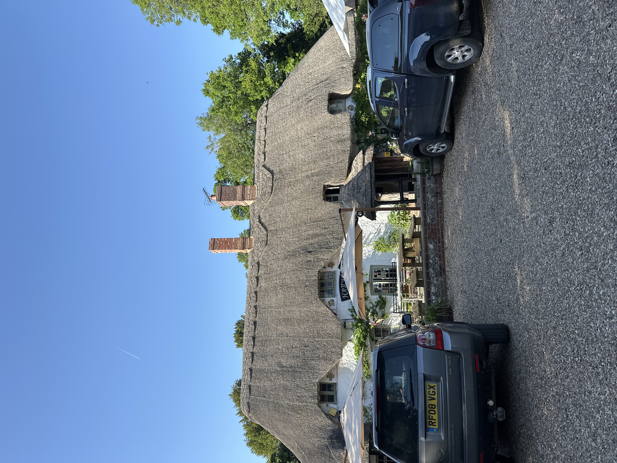 The image shows a thatched-roof building, possibly a pub or inn, with two cars parked nearby. The building has brick chimneys and several windows, with a sign partially visible that says "HOTEL". Outdoor seating is arranged near the building's entrance. The setting appears to be a rural or village location with gravel ground. The sky is clear and blue. The image is taken during daylight hours. A license plate on one car reads "RF08 VGX". The location is in the city of Unknown, Unknown.