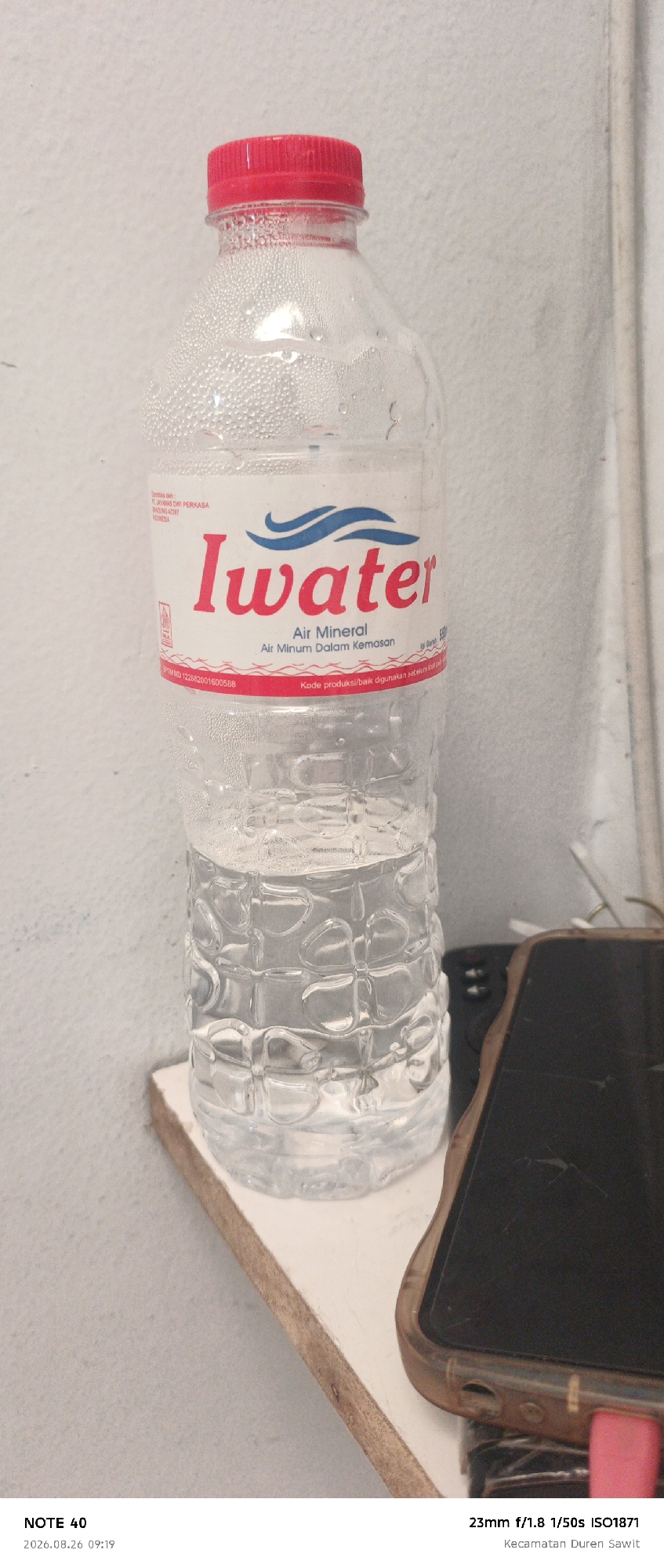 The image shows a clear plastic bottle of "Iwater" brand mineral water with a red cap, placed on a light-colored surface. Condensation is visible on the outside of the bottle. In the background, there is a lightly textured wall. To the right of the water bottle, part of a dark-colored smartphone with a brown case is visible. The bottom of the image contains text indicating "NOTE 40", a date and time "2026.08.26 09:19", camera settings "23mm f/1.8 1/50s ISO1871", and a location "Kecamatan Duren Sawit". The scene appears to be indoors.