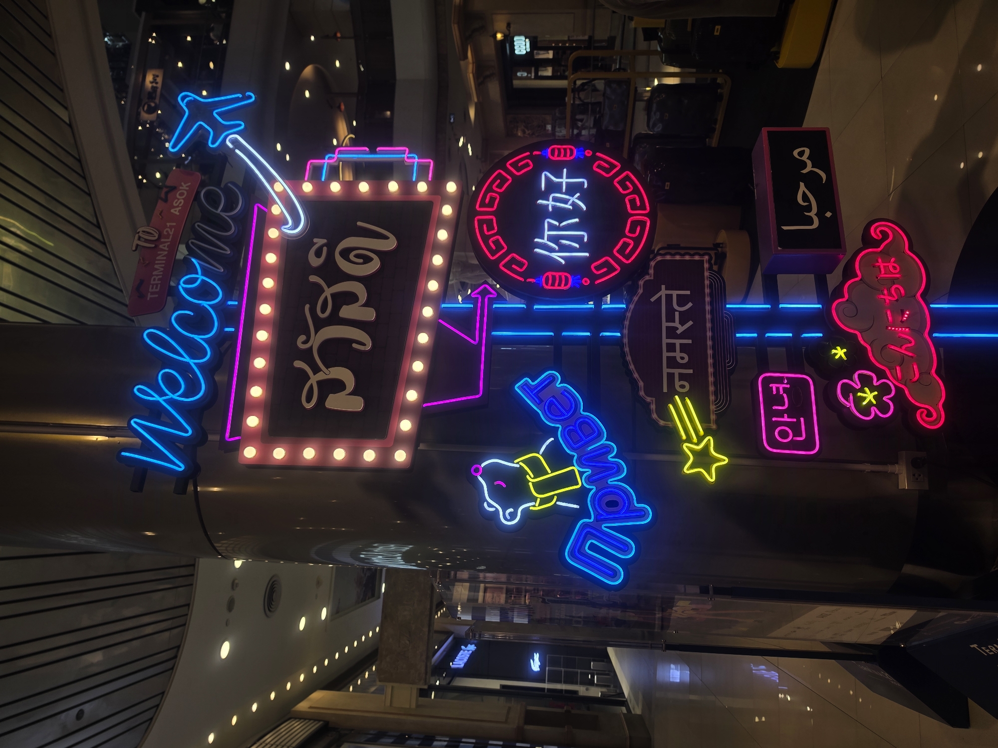 The image shows a collection of neon signs in various languages, including English ("Welcome to Terminal 21 Asok"), Thai ("สวัสดี"), Chinese ("你好"), Hindi ("नमस्ते"), Korean ("안녕"), and Arabic ("مرحباً"). These signs are likely part of the decorations inside the Terminal 21 Asok shopping mall in Bangkok, Thailand. There is a neon airplane above the "Welcome" sign and other neon figures such as a bear. The signs are mounted on a pillar. The lighting is dim, highlighting the neon signs. The background shows elements of the shopping mall's interior, with various shops and structural supports.
