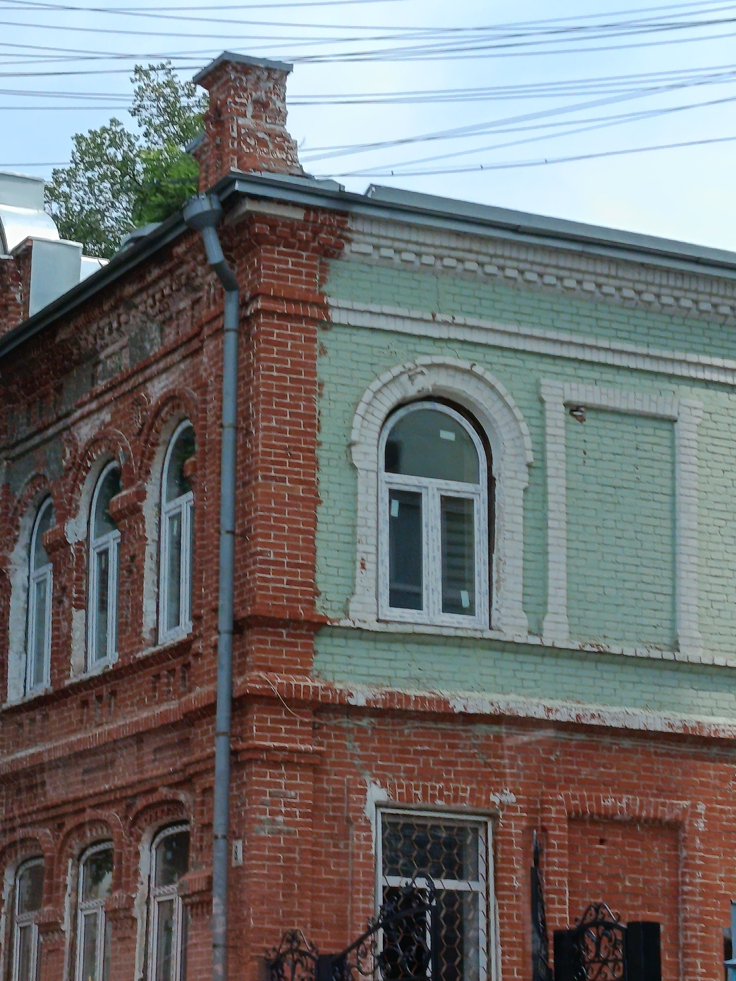 The image shows a close-up of the upper corner of a red brick building with a green painted section on the right. The building features multiple arched windows on the red brick facade, some with white frames and glass panes. One large arched window on the green section also has a white frame and glass. A metal downspout runs vertically along the corner of the building. Above the building, power lines crisscross the sky, and a tree with green foliage is visible in the upper left corner. The architecture suggests an older building, possibly from the late 19th or early 20th century. The specific location in Saratov, Russia, is not discernible from the image itself.