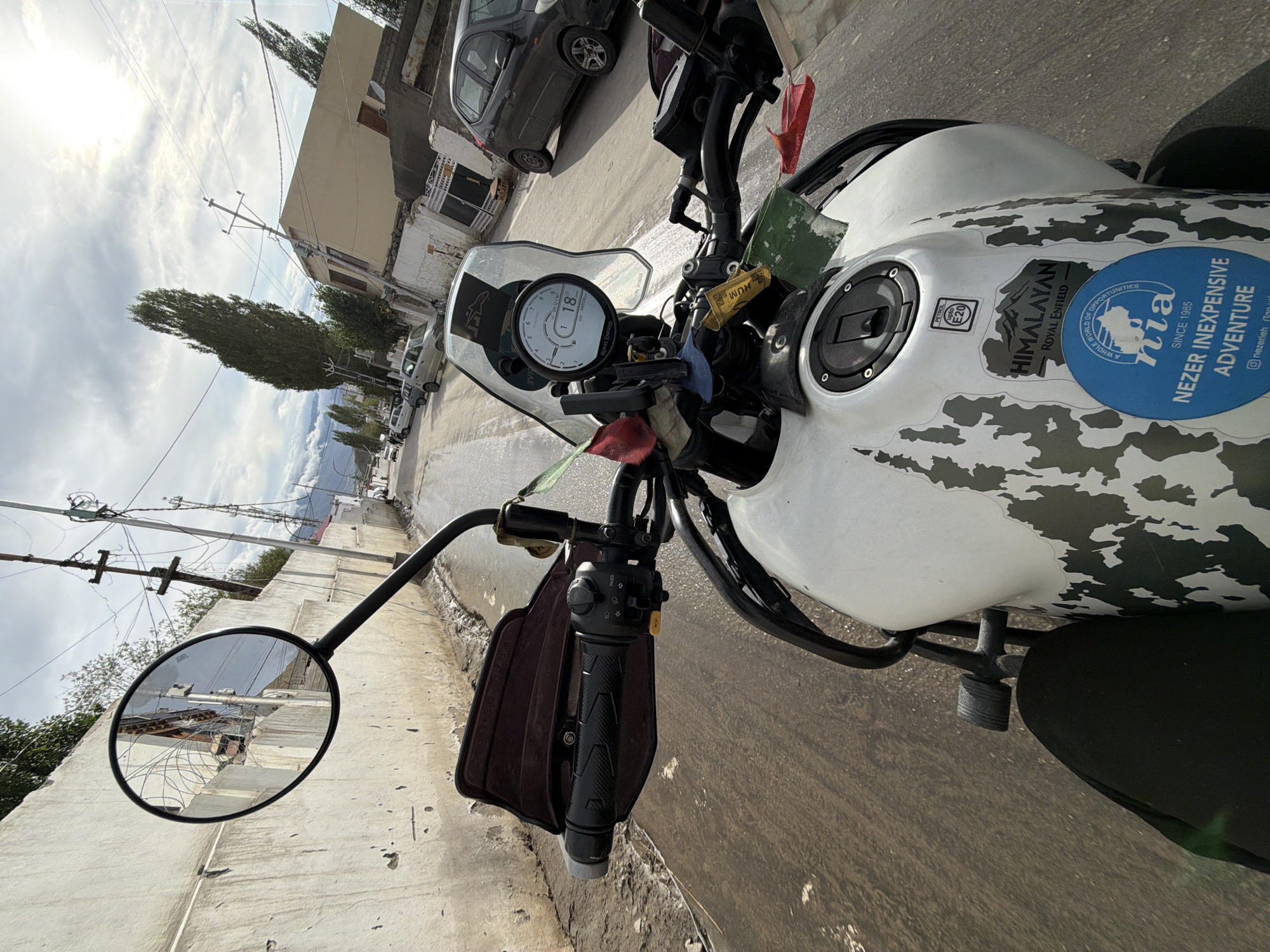The image is a first-person perspective from a motorcycle in Leh, India. The motorcycle is white with a camouflage-like pattern and has several stickers on its fuel tank, including one that reads "HIMALAYAN ROYAL ENFIELD" and another blue circular sticker with text. The rider's black pant leg is visible in the lower left corner. The motorcycle's handlebars, mirrors, and digital display are in the foreground. The display shows the speed as 18 km/h and the time as 05:12. Prayer flags are attached to the motorcycle. The road appears wet and somewhat unpaved, with a concrete wall on the left side. Buildings, utility poles with wires, and parked cars are visible along the street, suggesting an urban or semi-urban environment. The sky is cloudy with the sun peeking through. The overall scene depicts a motorcycle ride through a town in a mountainous region.