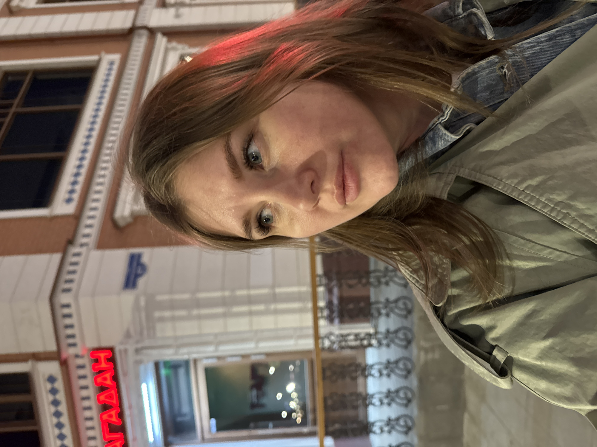 A woman is photographed on a street in Perm, Russia, identifiable by the "АГАААН" sign displayed on a building behind her. She has light brown hair with some red highlights and is wearing a light green coat over a denim jacket. The building features windows and patterned architectural details. The street is visible in the background, with decorative metalwork and other architectural features. The photo appears to have been taken in the evening or night, as indicated by the lighting.