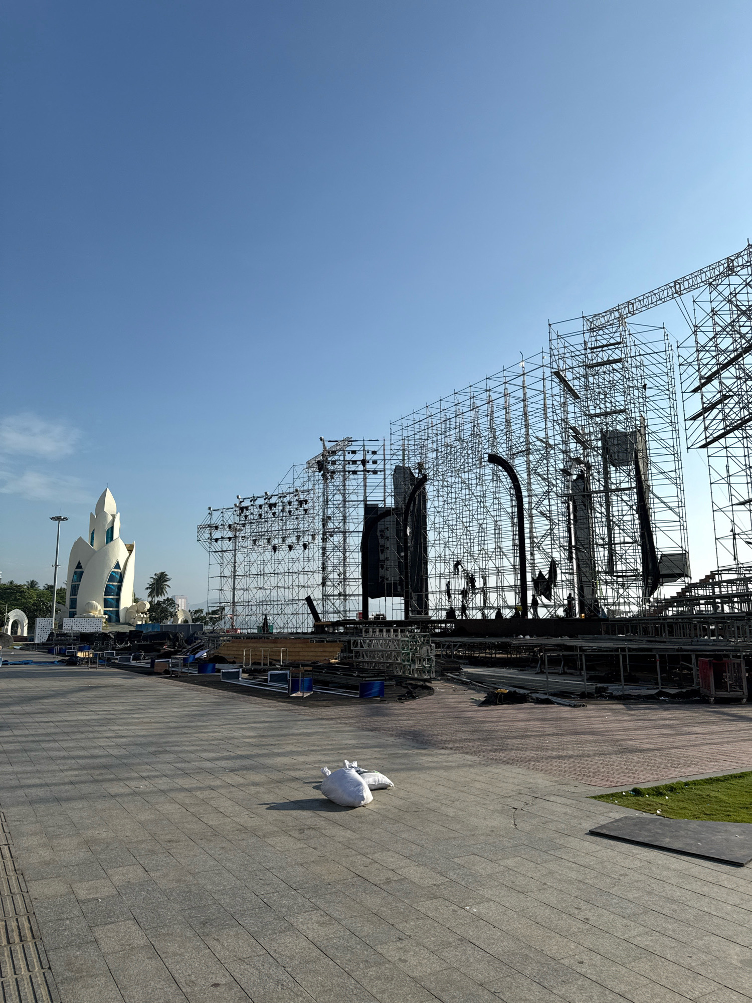 The image depicts a large outdoor construction site, likely a stage being erected for an event, located in the city of Nha Trang, Vietnam. In the background, a distinctive white building with a unique, petal-like structure stands tall against a clear blue sky. To the right of the white building, an extensive scaffolding system dominates the scene, suggesting the construction of a large-scale stage or display. Several figures are visible on the scaffolding, appearing to be workers engaged in assembly or maintenance.
The foreground of the image shows a paved plaza area with stone tiles. Two white sacks, possibly filled with sand or other materials, are placed on the pavement. Beyond the plaza, to the right, a neatly trimmed patch of green grass is visible. The overall impression is one of ongoing preparation for a significant event in a coastal city. The architecture of the white building and the presence of palm trees in the distance further indicate a tropical or subtropical location.