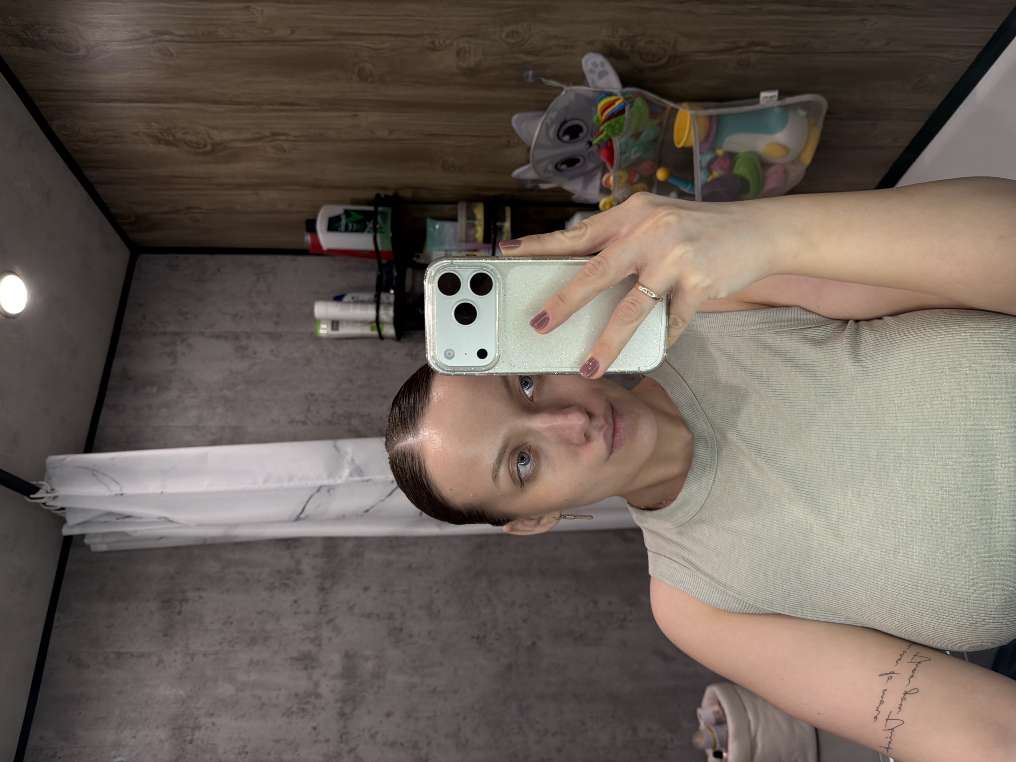 An individual is captured taking a mirror selfie in what appears to be a compact bathroom setting in Reshetnikovo, Russia. The image is oriented vertically, with the person positioned on the right side.
The subject is a person with dark hair slicked back, revealing a clear complexion and light-colored eyes. She wears a light green or tan ribbed tank top and a gold-colored ring on her right ring finger. A subtle gold chain-link earring is visible on her left ear. On her right forearm, a tattoo displays two lines of script text, primarily illegible but possibly in English, with phrases like "Live your dream" or "Love you more" suggested.
She holds a smartphone, likely an iPhone due to its distinct camera module, encased in a clear, sparkly cover. The phone partially obscures her face as she aims it towards the mirror to take the photo, with her gaze directed towards the camera.
The background shows the interior space. The walls are a dark grey with a textured, possibly concrete or tiled, finish. The ceiling or upper wall is clad with horizontal wooden planks. On the left, a white shower curtain with a grey marble-like pattern is visible, drawn along a corner. A round, illuminated light fixture is present on the left wall. Behind the phone and to the right, a collection of various bottles and containers, presumably personal care products, are arranged on a shelf or ledge. Above these items, a grey fabric cat-shaped toy with colorful attachments and a mesh bag filled with colorful baby toys, including a yellow cup and a blue scoop, are visible. The lighting appears to be artificial, typical of an indoor environment.